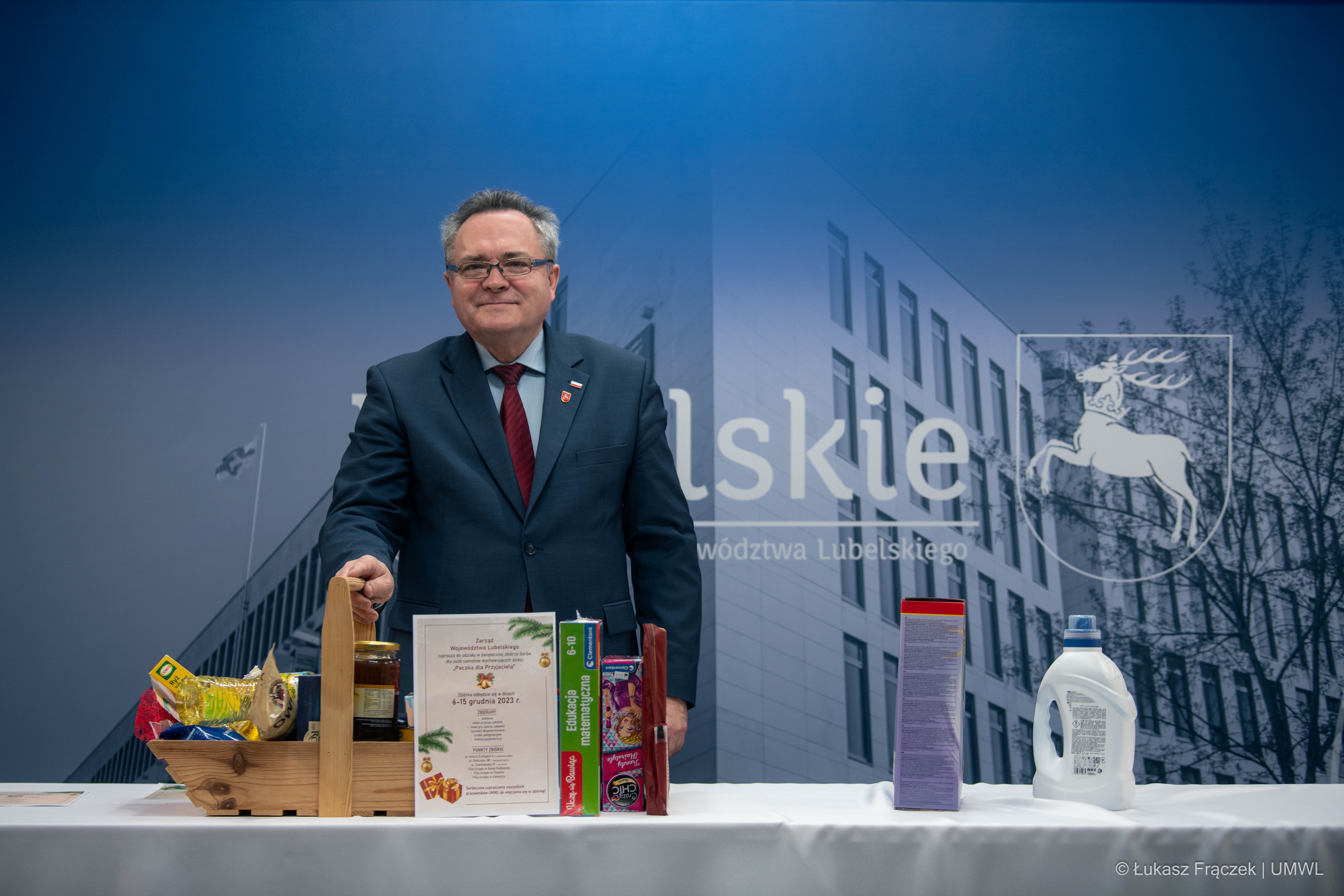 Wicemarszałek Zbigniew Wojciechowski stoi przy stole na tle ścianki lubelskie a przed nim na stoliku w koszu stoją produkty spożywcze