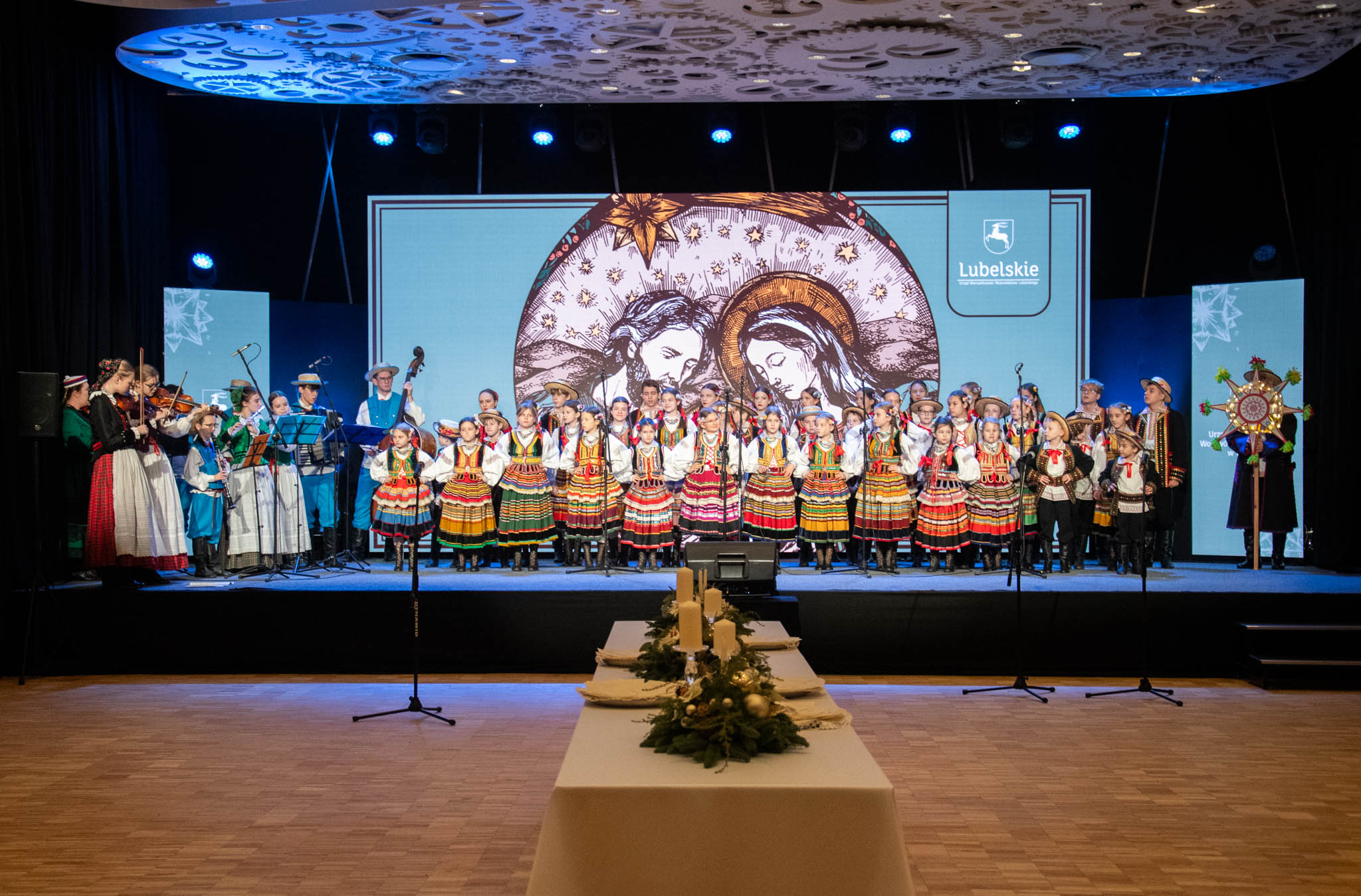 widok na scene na której stoi dziecięcy zespół ludowy, łacznie około dwudziestu dzieci w strojach ludowych, przed nimi na sali stoi stół udekorowany świerkiem i świecami