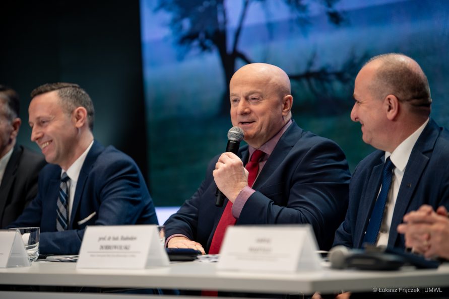 Marszałek Jarosław Stawiarski siedzi przy stole konferencyjnym w towarzystwie pozostałych uczestników udziela wypowiedzi