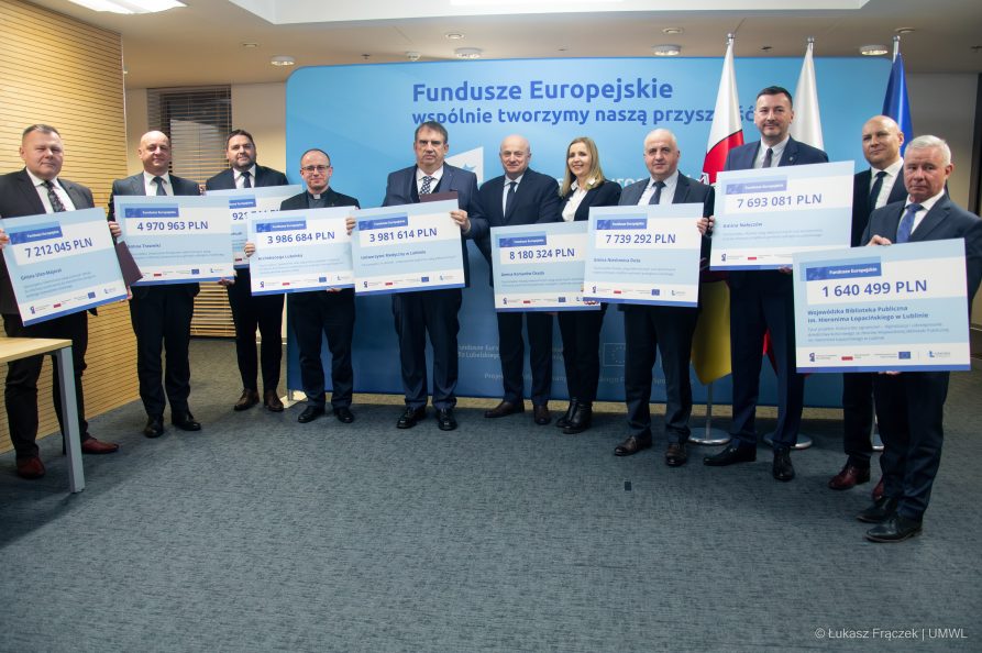 Około dziesięciu osób, które stoją wspólnie w sali konferencyjnej i trzymaja przed sobą symboliczne tabliczki