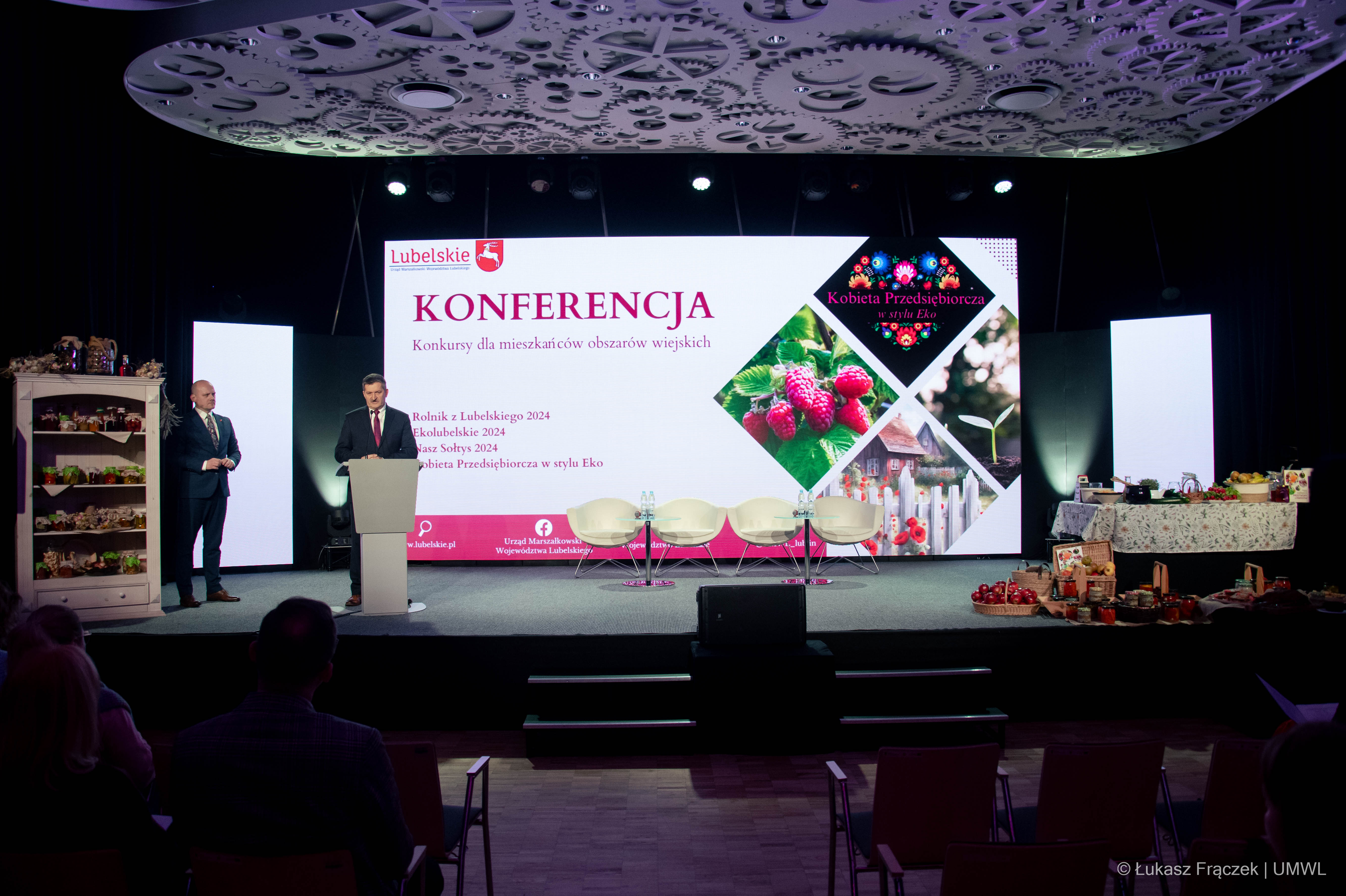 Zdzisław Szwed udziela wypowiedzi do mikrofonu, za nim znajduje się duży ekran wyświetlający napis Konferencja