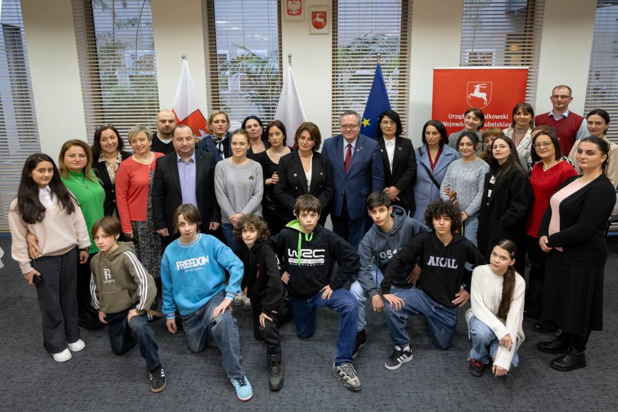 Grupa gości z Gurii pozująca na tle flag wraz z Wicemarszałkiem Zbigniewem Wojciechowskim