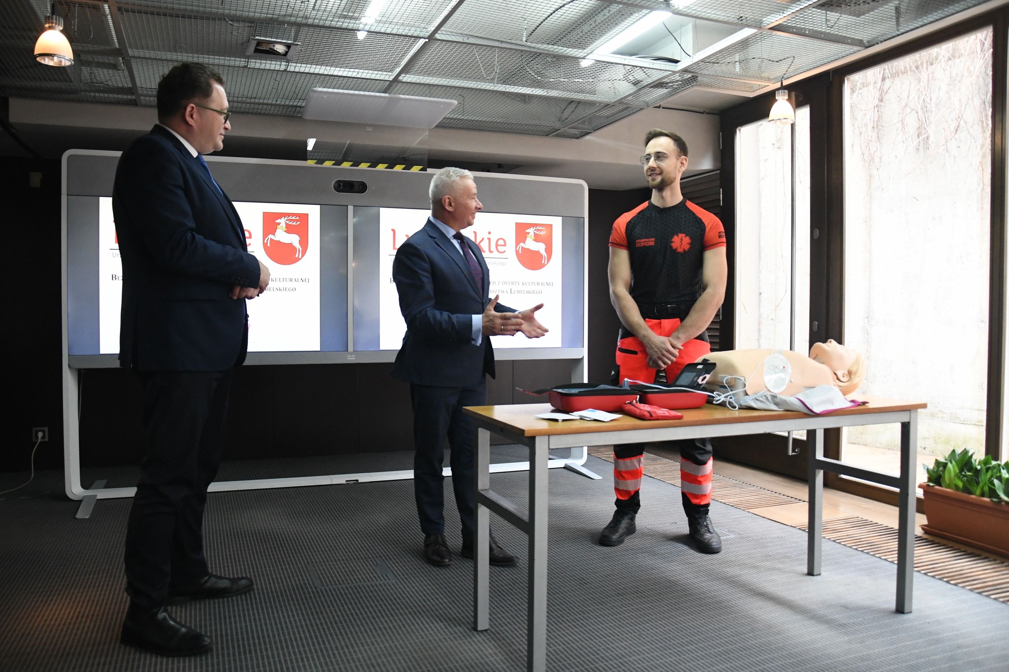 Członek Zarządu Bartłomiej Bałaban i Dyrektor Bilbioteki przyglądająsię pracy ratownika medycznego, który pokazuje jak korzystać z defibrylatora na manekinie