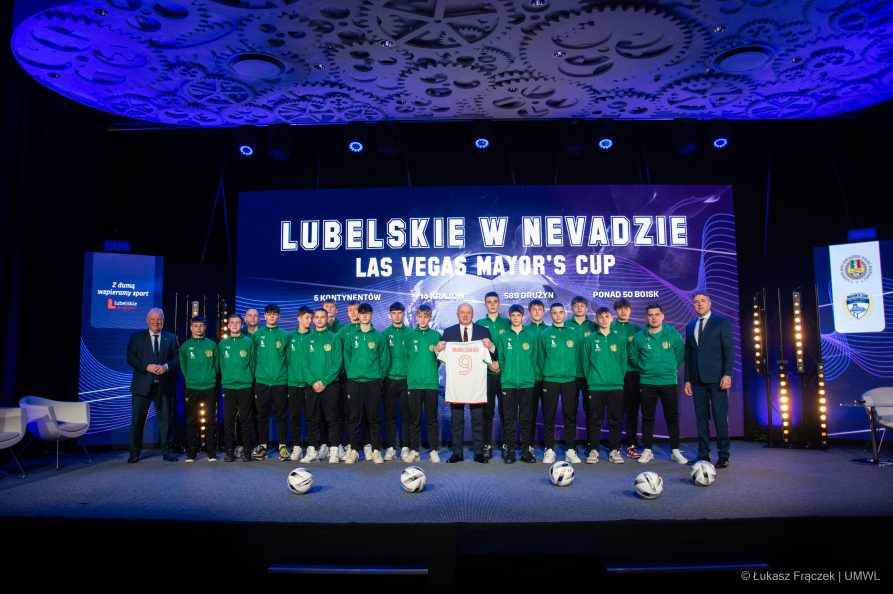 Marszałek WL trzyma w rekach koszulke sportową i pokazuje ją do kamery. Obok niego stoją członkowie piłkarskiej drużyny chłopców