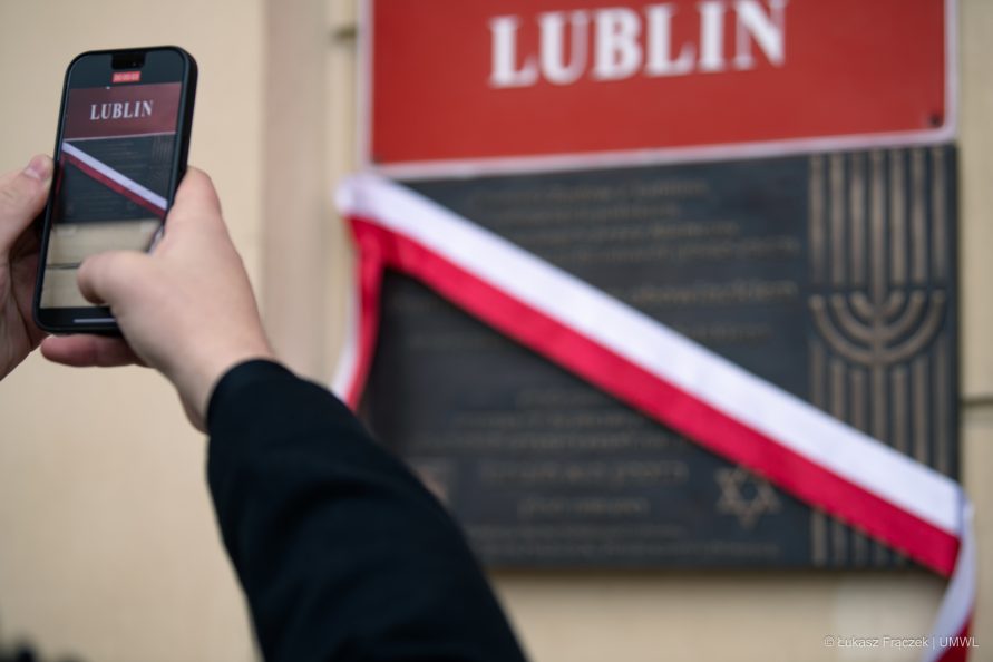 Widok na dłonie w których trzymany jest telefon. Właściciel telefonu robi zdjęcie tablicy wiszącej na ścianie urzędu