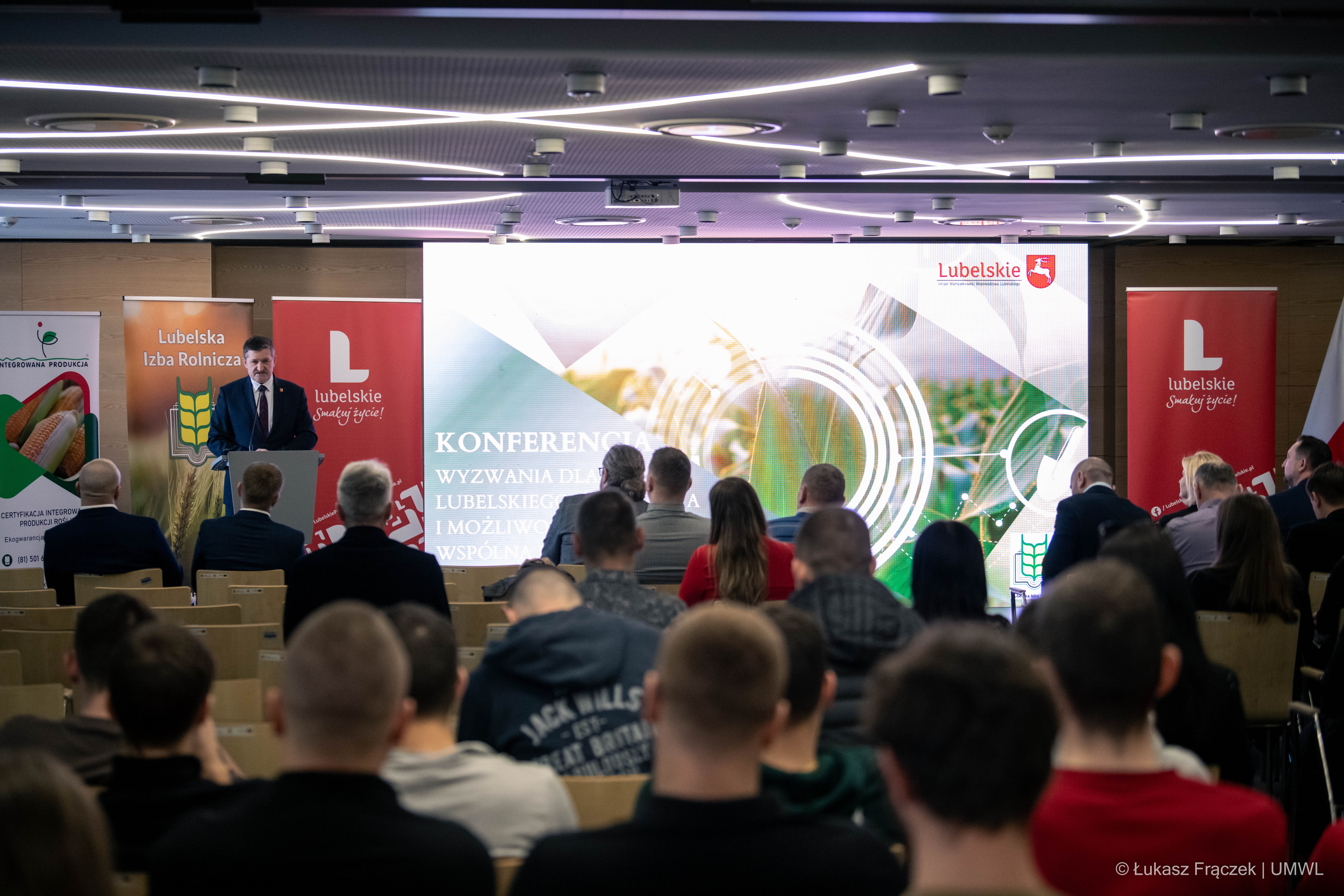 Zdzisław Szwed stoi przy mównicy i przemawia. Na pierwszym planie widać kilkadziesiat osób siedzacych i słuchających konferencji. Na środku sali znajduje się ekran wyświetlający grafiki o tresci rolniczej