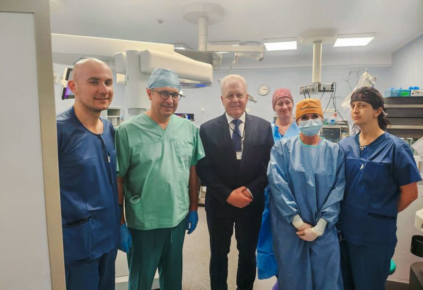 Zespół lekarzy chirurgów ubranych w kitle stoją na sali operacyjnej. Między nimi stoi ubrany w garnitur dyrektor szpitala