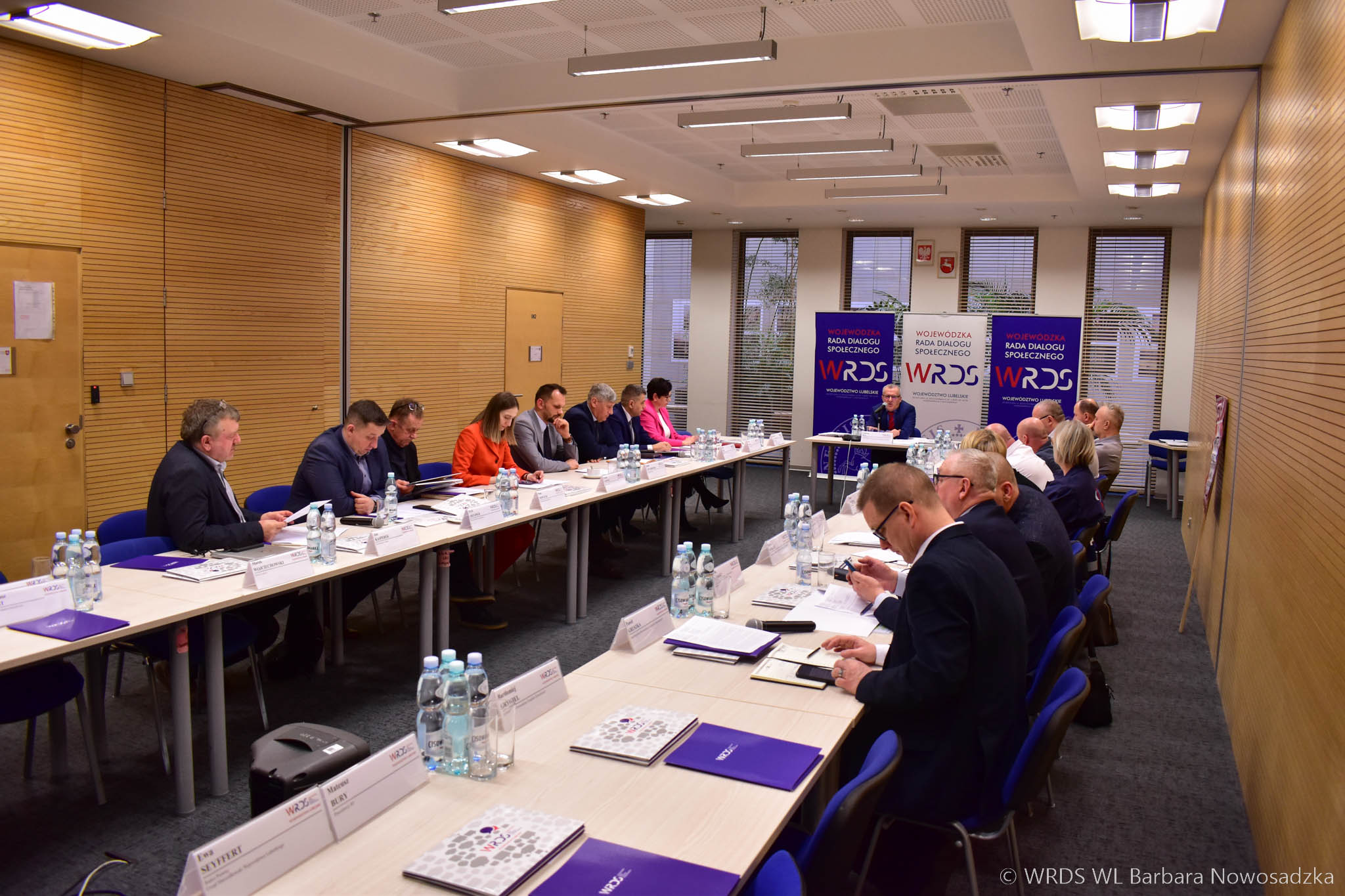 Sala konferencyjna na której znajduje się długi stół ustawiony w podkowę i siedzi przy nim około dwudziestu osób, które uczestniczą w posiedzeniu i zaglądają w notatki