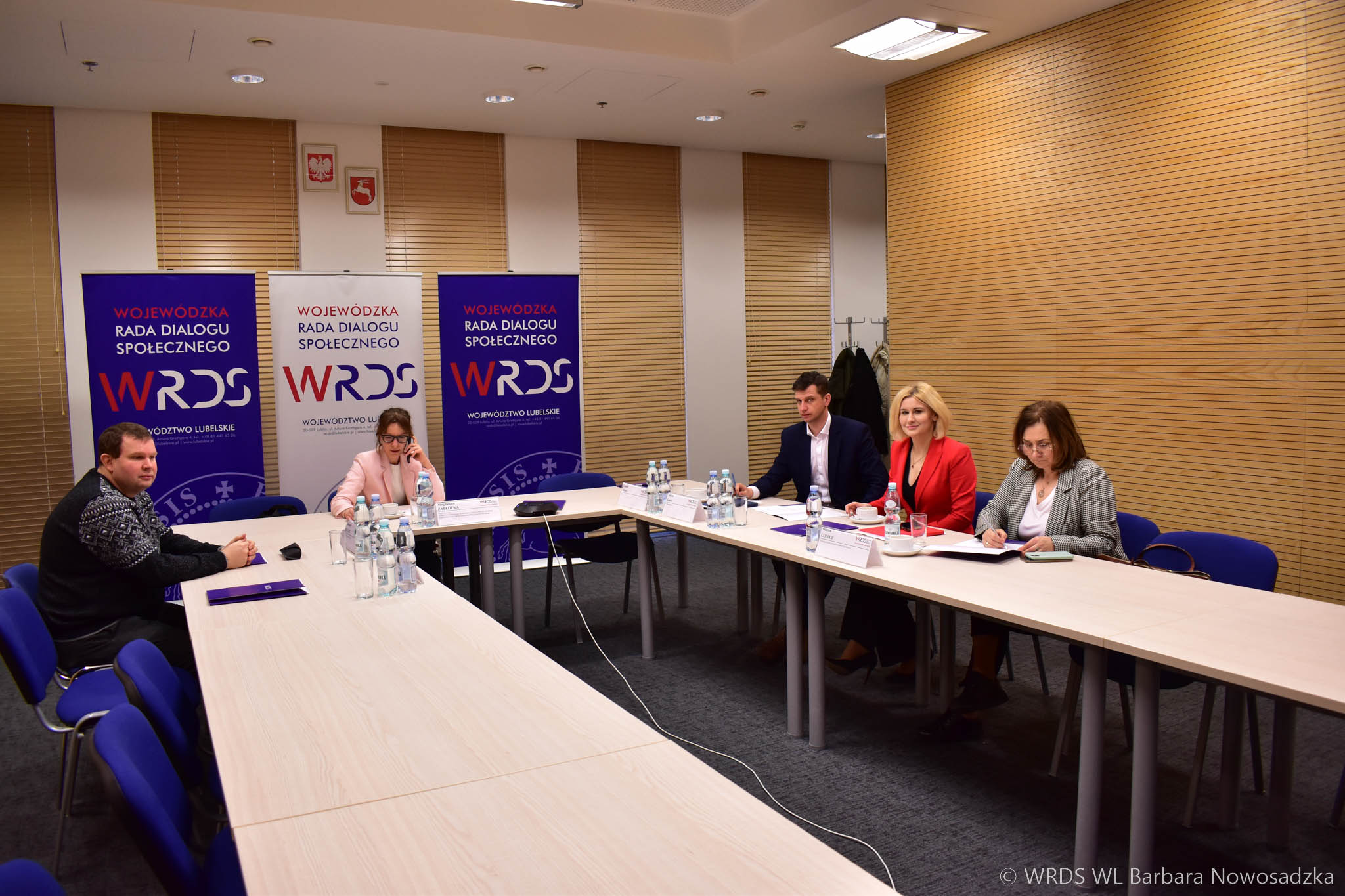 Wnętrze sali konferencyjnej w której są ustawione stoły w podkowę przy których siedzą trzy osoby i jeden mężczyzna.