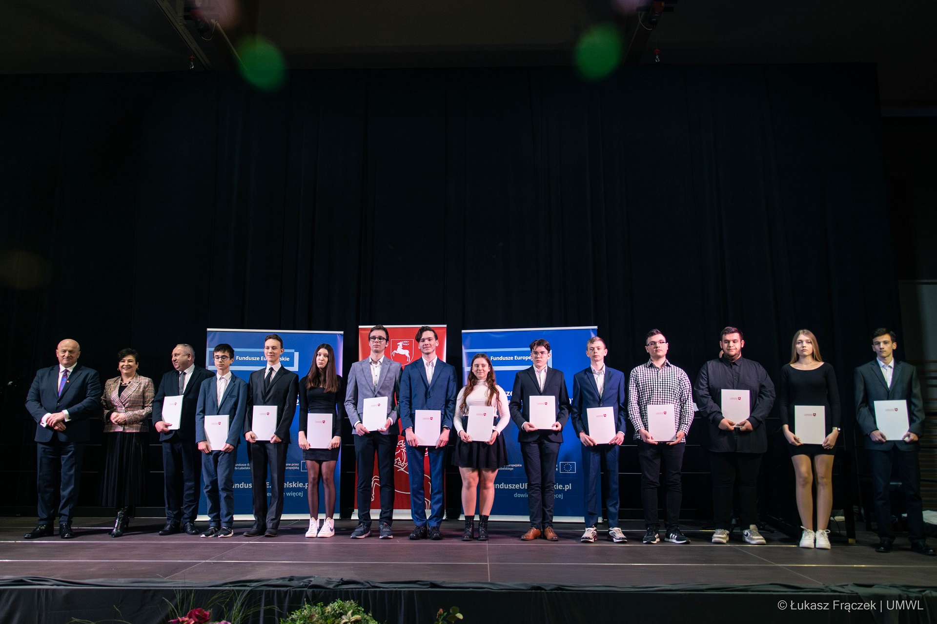 Około kilkunastu uczniów, którzy otrzymali stypendia, stoja na scenie i pozują do zdjęcia, każdy z nich trzyma w ręku teczkę z otrzymanym stypendium