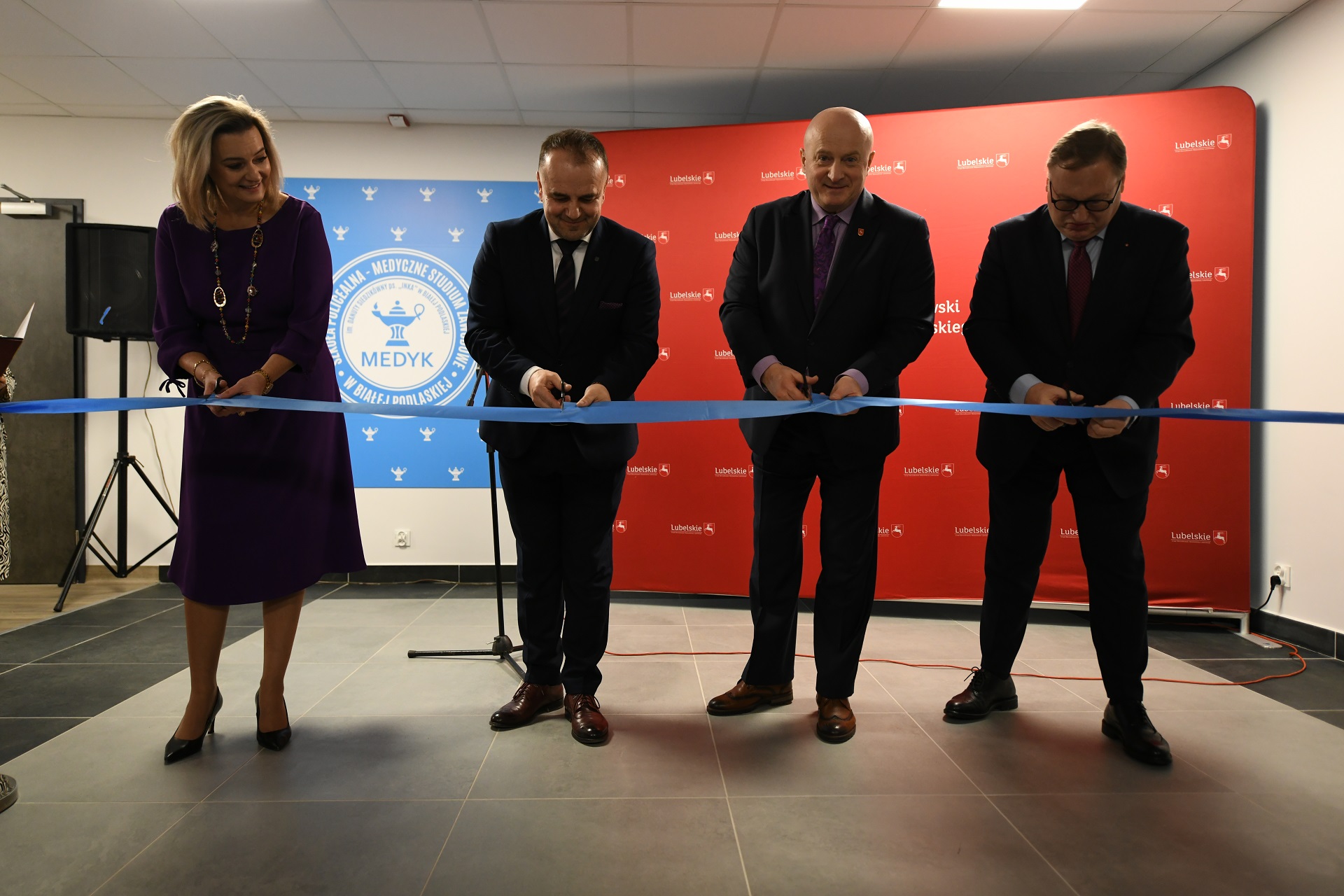 Trzech mężczyzn i jedna kobieta stoja we wnętrzu pomieszczenia i przecinają wstęgę