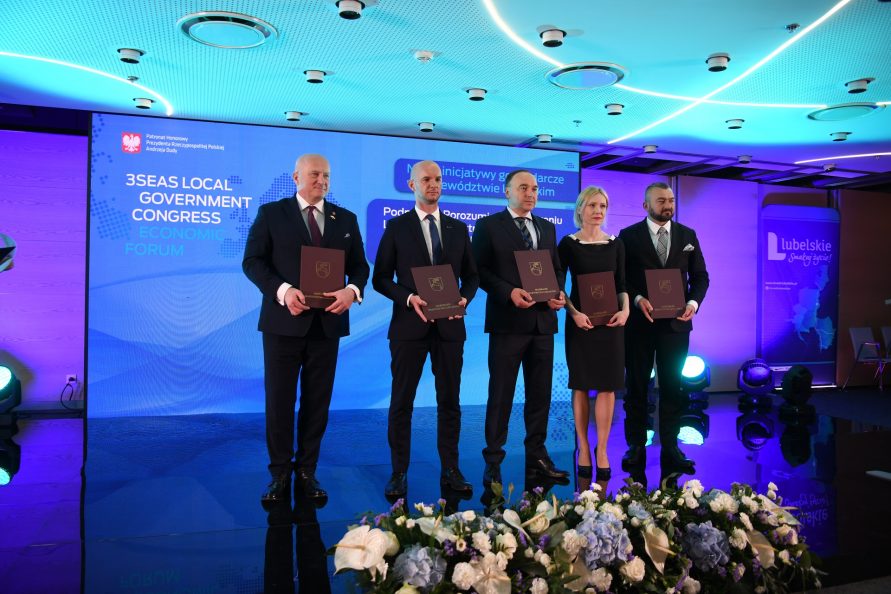 Marszałek Województwa Lubelskiego wraz z kilkoma pozostałymi uczestnikami podpisania umowy, wszyscy wspólnie stoją na podeście i prezentują teczki z podpisanymi umowami