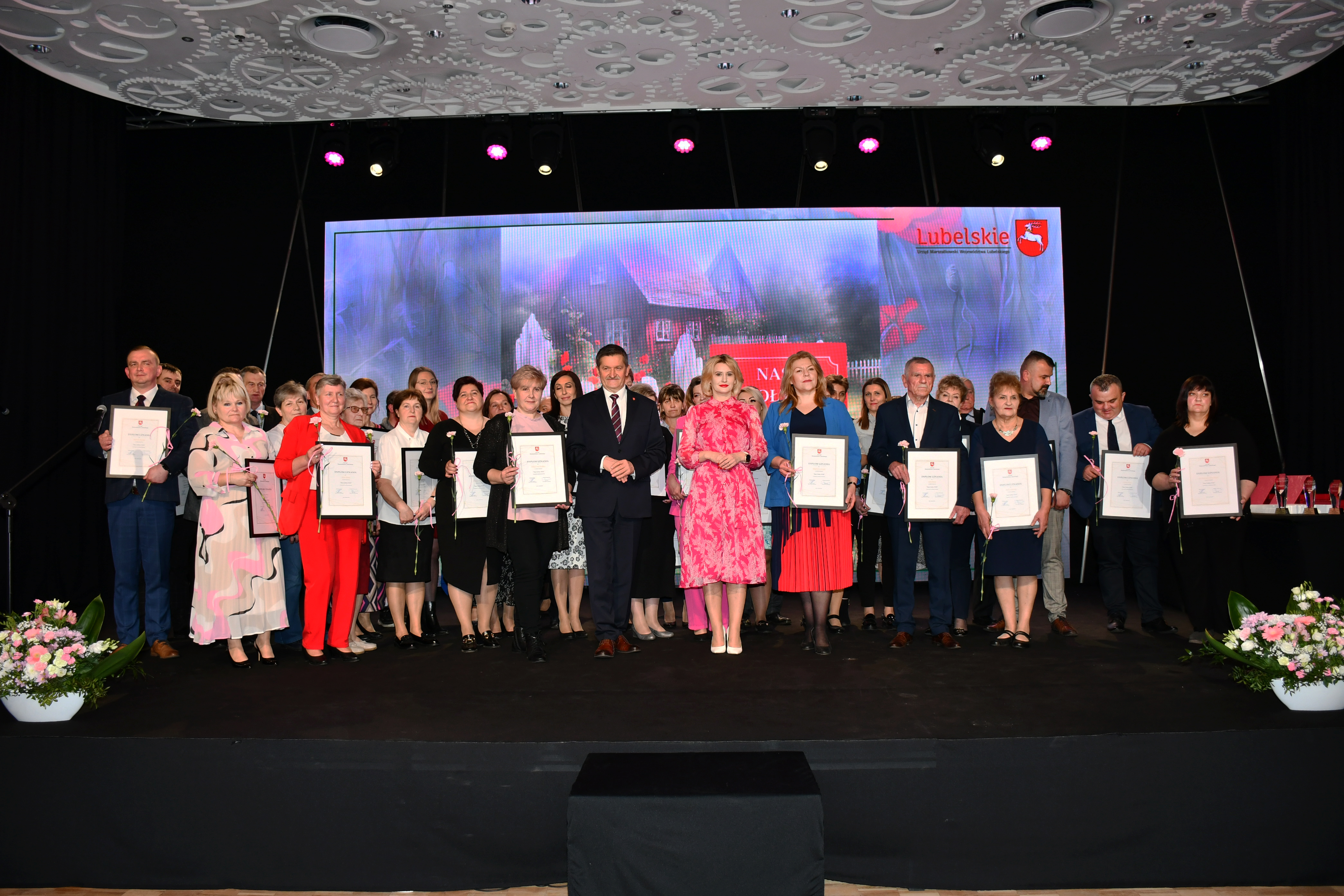 Około dwudziestu osób stoi na scenie i pozuje do zdjecia. Są to laureaci i wyróżnienie w konkursie nasz sołtys w towarzystwie członka zarządu