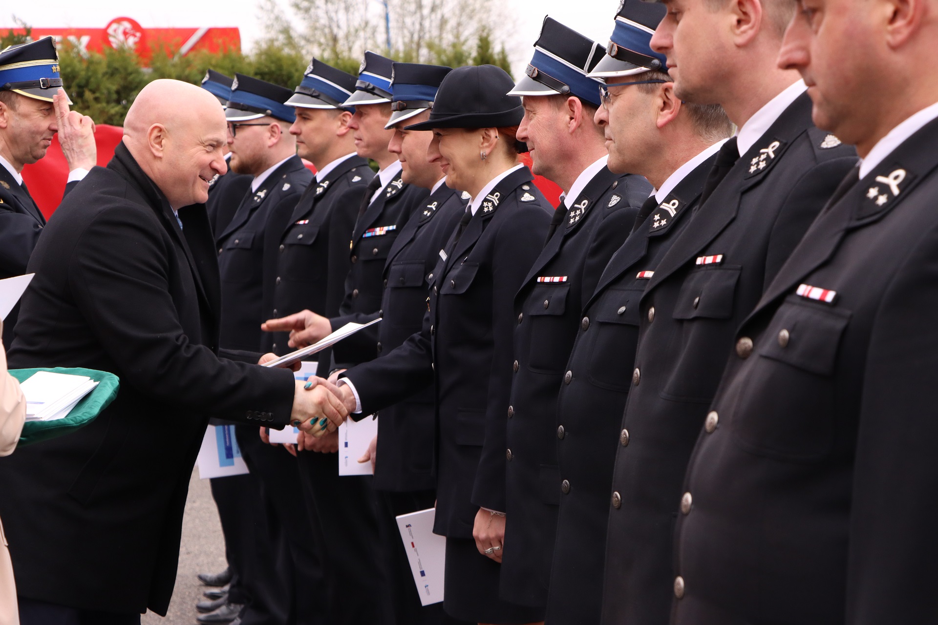 Marszałek Jarosław Stawiarski podaje rękę kobiecie strażaczce. Dookoła stoją strażacy, około dziesięciu osób w galowych mundurach