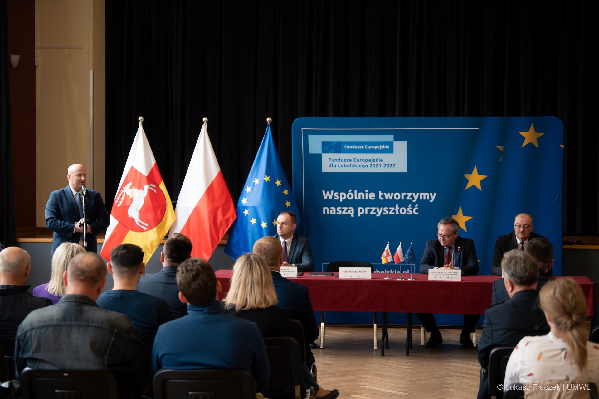 Marszałek Jarosław Stawiarski stoi przy mównicy i przemawia do zebranych. Na pierwszym planie widać około dziesięciu osób siedzących na widowni. Obok marszałka stoją trzy flagi a dalej przy długim stole siedzi trzech mężczyzn.