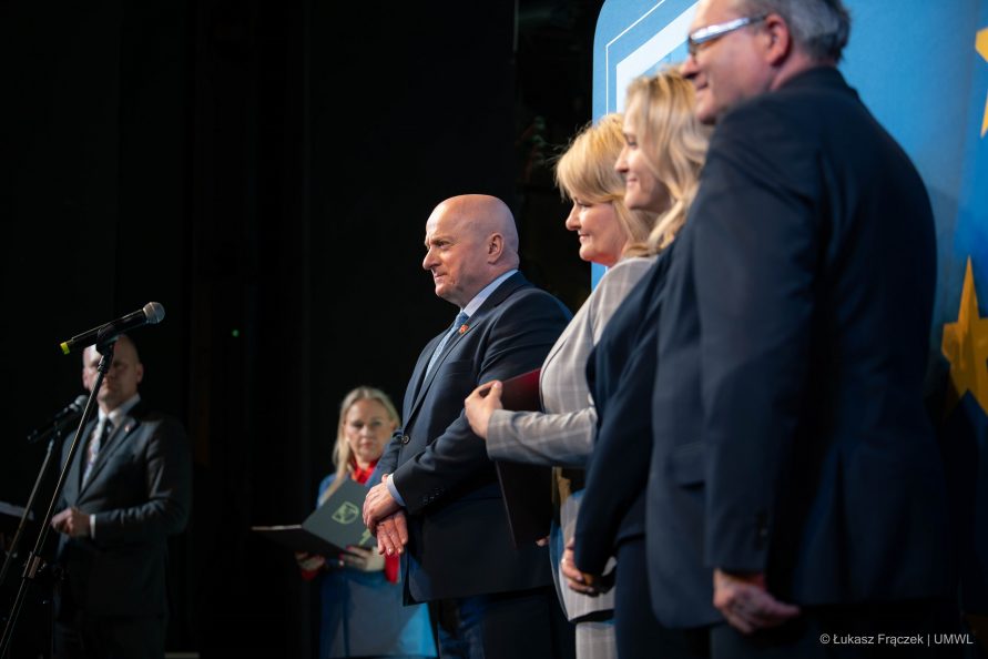 Pięć osób stoi na scenie w ujęciu profilowym. Wśród nich znajduje się marszałek Jarosław Stawiarski
