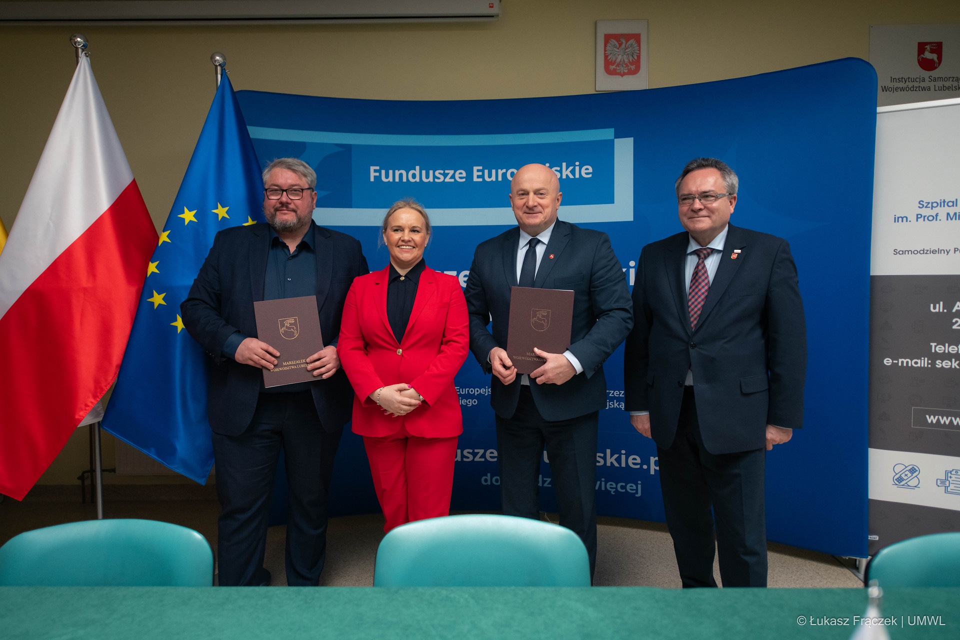 Trzech mężczyzn i jedna kobieta stoją w sali konferencyjnej i pozują do zdjęcia . Za nimi znajduje się ścianka promująca fundusze europejskie