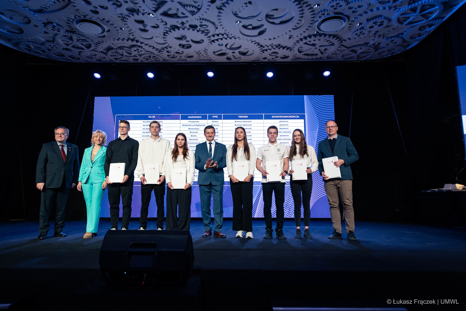 Na scenie stoi dwóch członków zarządu oraz dyrektorka z UMWL którzy towarzyszą kilku młodym ludziom, odbierającym sportowe stypendia marszałka