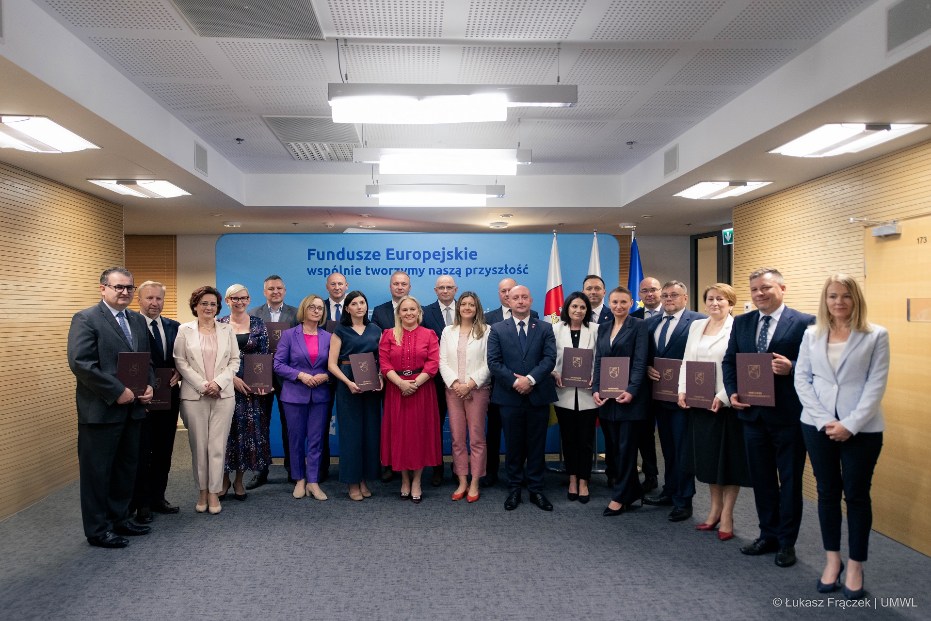 Grupa około dwudziestu osób pozuje wspólnie do zdjęcia. Wszyscy stoją w sali konferencyjnej, niektórzy z nich prezentują przed soba teczki w których znajduje się otrzymany dokument zdofinansowaniem