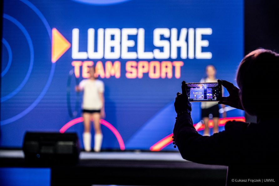 Duży ekran i napis na nim Lubelskie Team Sport. Na scenie przed ekranem stoi dwójka dzieci w wieku szkolnym ubranym w stroje gimanstyczne w trakcie pokazu gimnastyki
