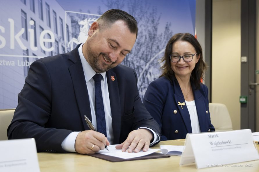 Wicemarszałek Marek Wojciechowski siedzi przy stole i podpisuje dokument, obok niego siędzi kobieta i uśmiecha się