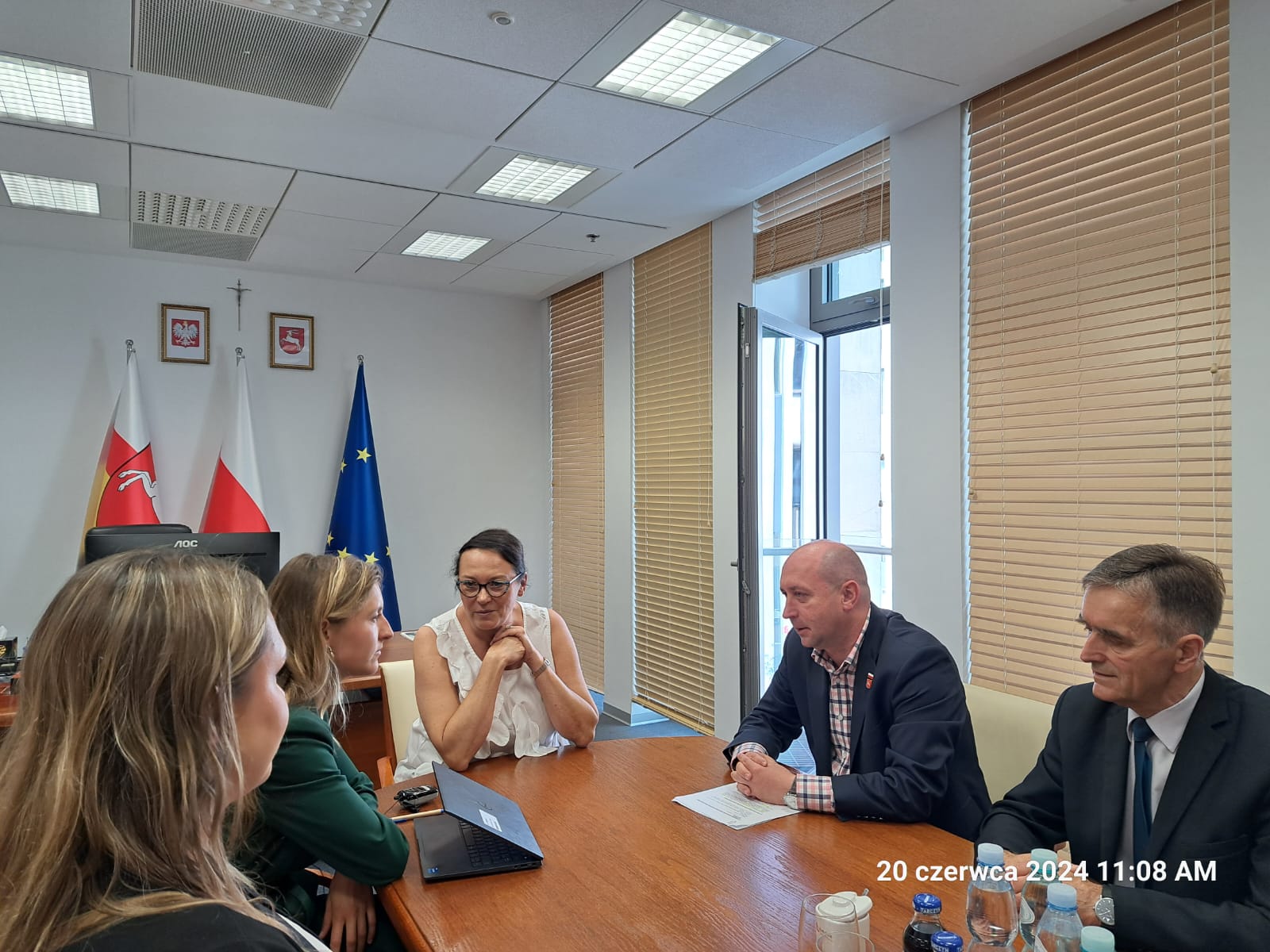 Przy stole siedzi dwóch mężczyzn i trzy kobiety, którzy wspólnie rozmawiają. W tle widać otwarte okno balkonowe w pokoju i stojące wewnątrz flagi Polski, województwa lubelskiego i UE