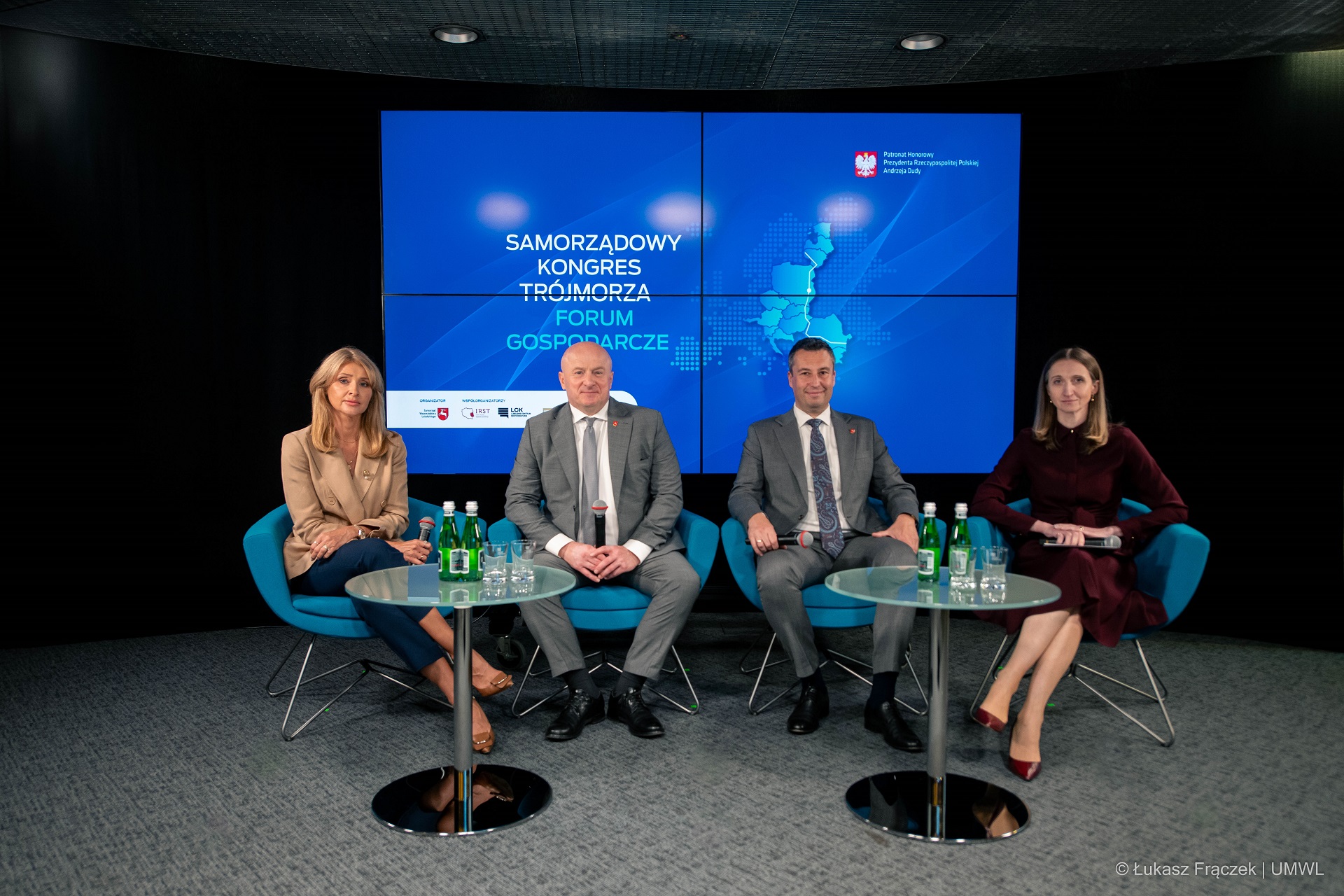 Dwie kobiety i dwóch mężczyzn siedzą na fotelach ustawionych na scenie. Przed nimi sa ustawione dwa szklane stoliki na których znajduje się woda. Za nimi na dużym ekranie wyświetla się grafika dotycząca Kongresu Trójmorza