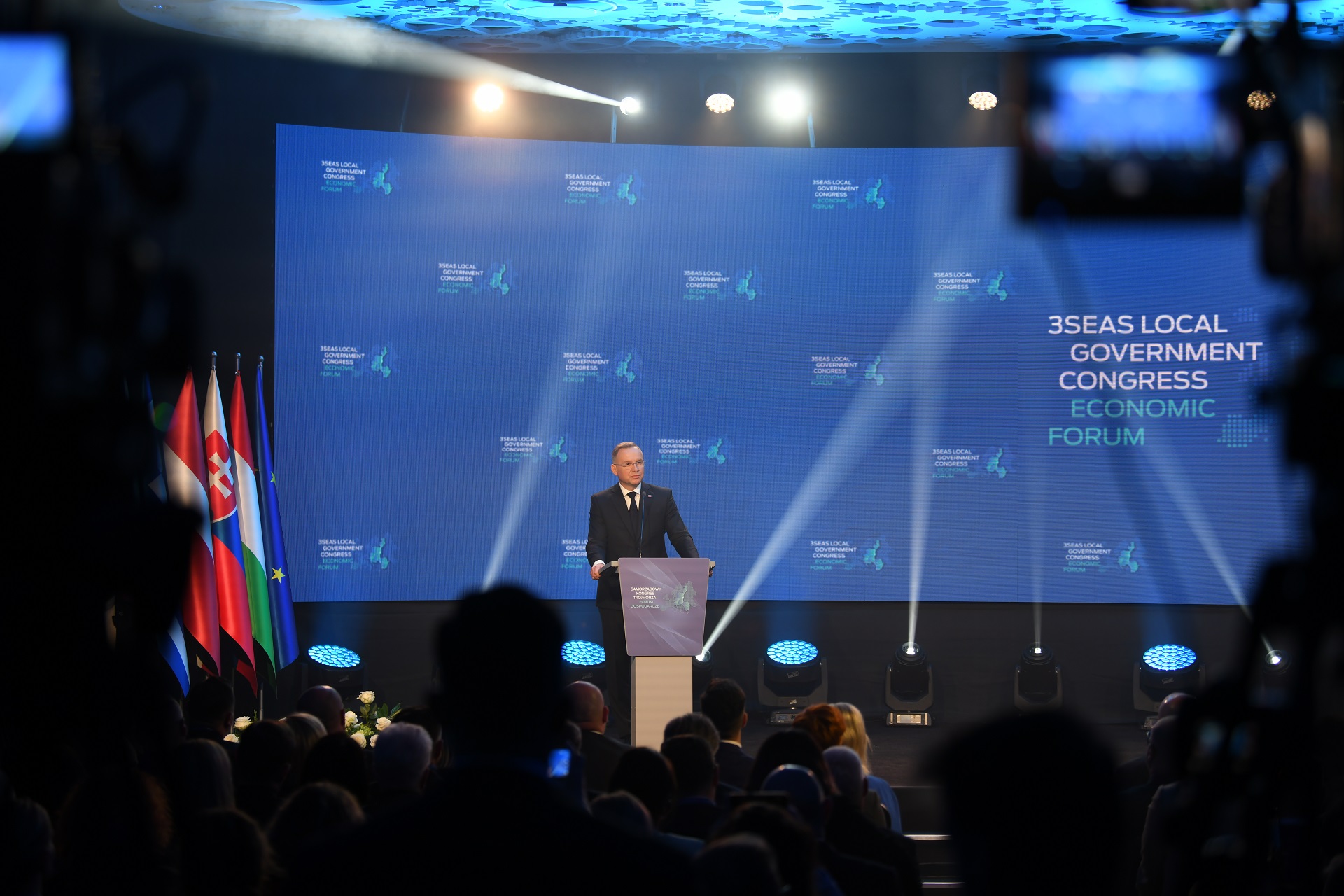 Prezydent Andrzej Duda stoi przy mównicy na scenie i przemawia do tłumu zebranego w sali konferencyjnej