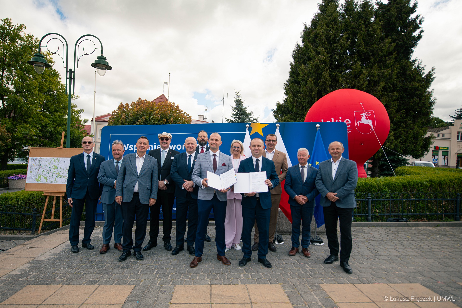 Około dziesięciu osób stoi na świeżym powietrzu na brukowanym placu. Wszyscy pozują do zdjęcia. Dwóch mężczyzn prezentuje otwarte teczki z umowami