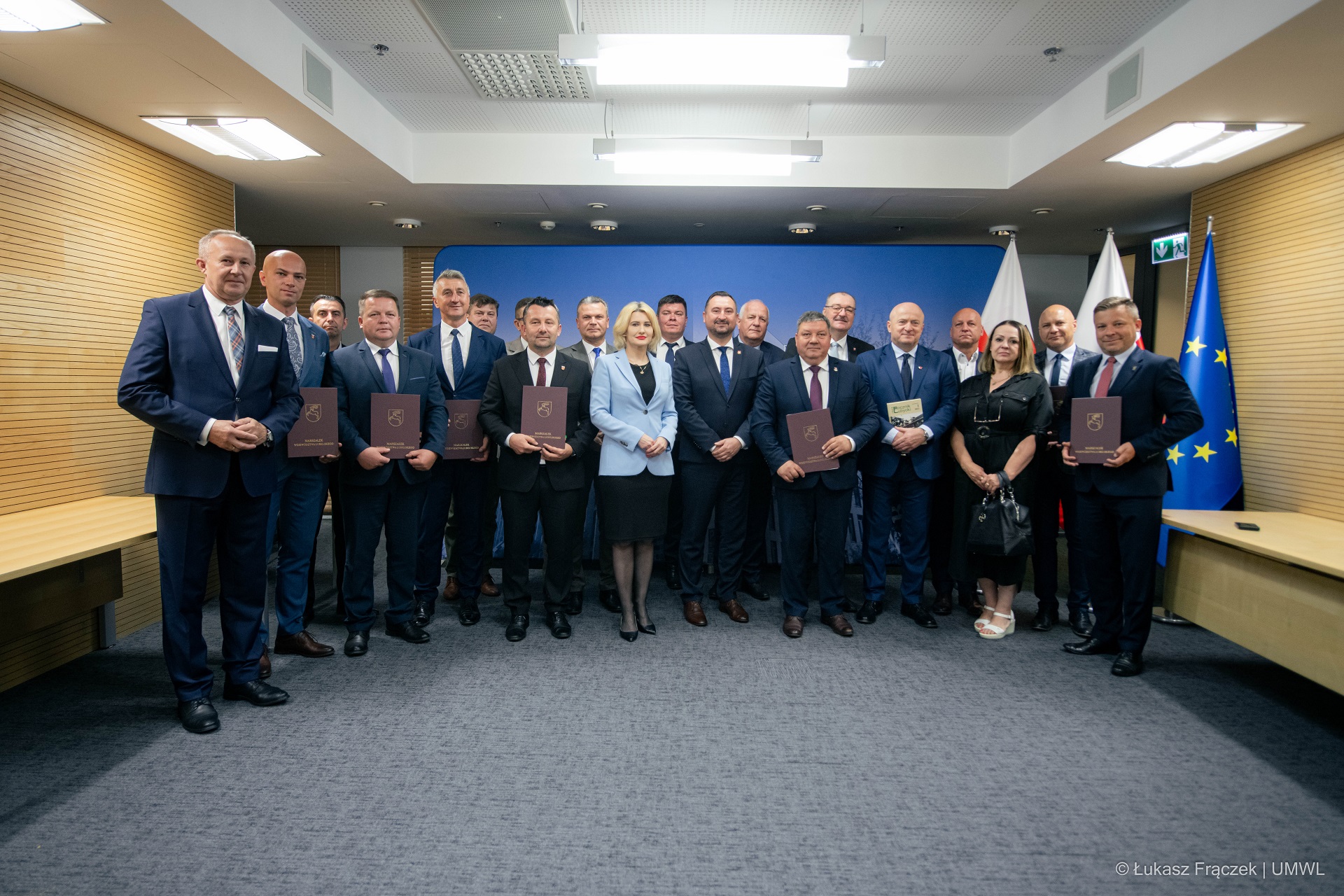 Na zdjęciu znajduje się kilkanaście osób. Wszyscy wspólnie pozują stojąc w sali konferencyjnej. Niektórzy z nich trzymają przed sobą teczki w których znajdują się podpisane umowy