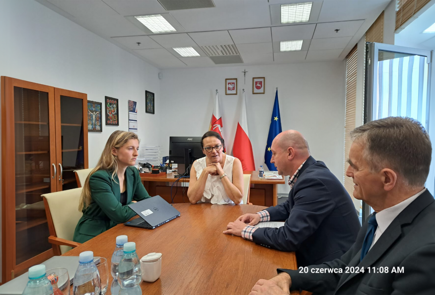 Przy stole siedzi dwóch mężczyzn i dwie kobiety, wszyscy ze sobą rozmawiają. Jedna z kobiet trzyma przed soba laptop. W tle przy ścianie pokoju widać stojące flagi Polski, województwa lubelskiego i UE