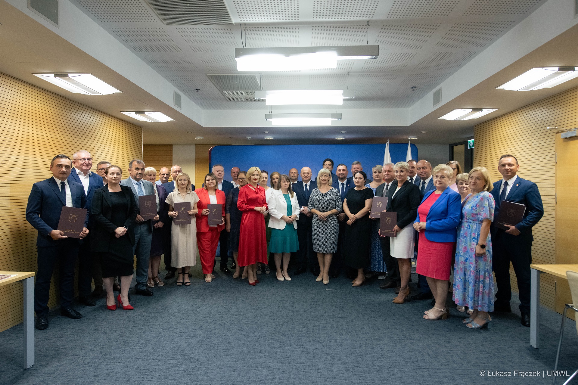 Około trzydziestu osób stoi w sali konferencyjnej w półkolu i wspólnie pozują do zdjęcia, są to przedstawiciele samorządów, którzy dostali dofinansowanie do budowy dróg
