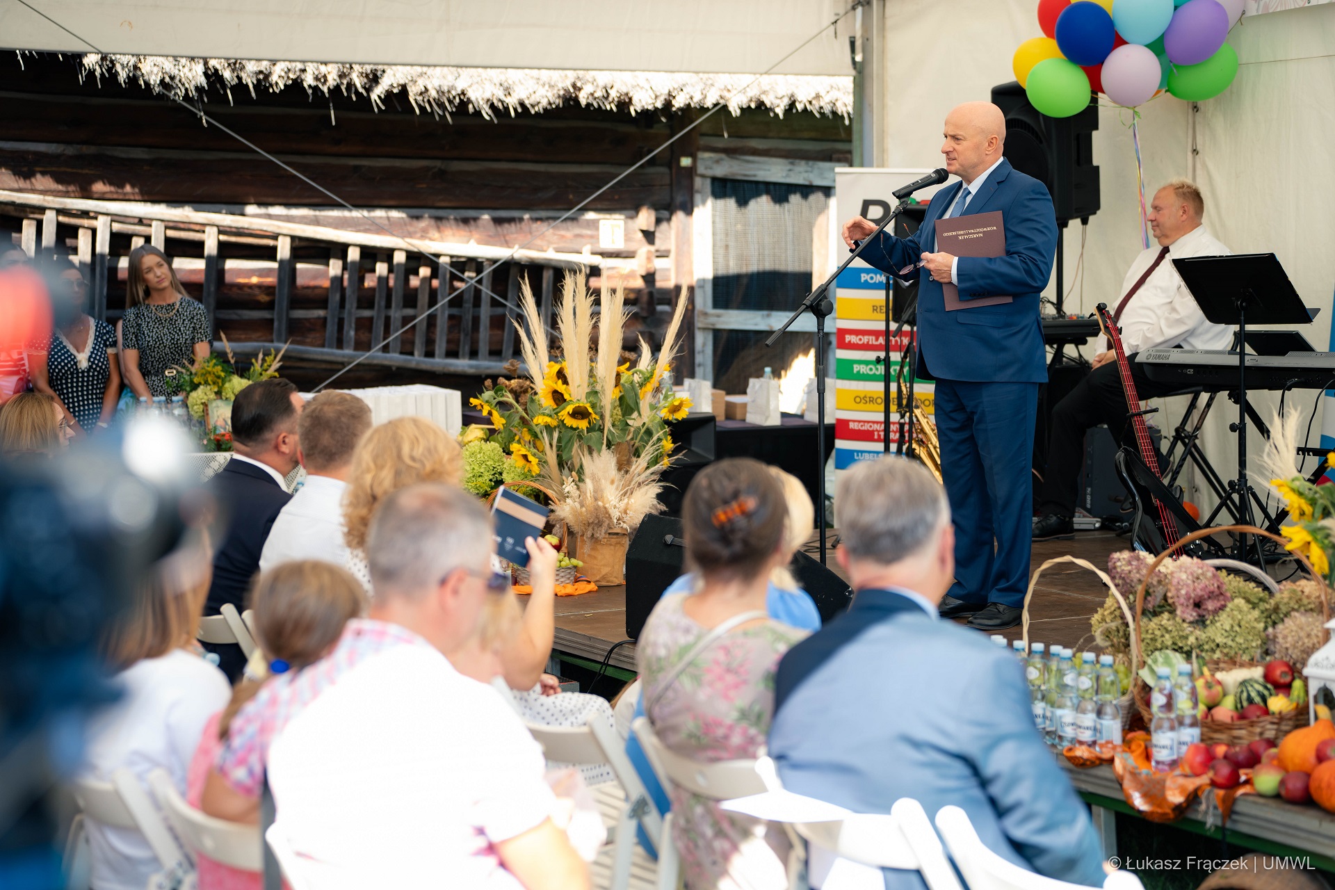 Marszałek Jarosław Stawiarski stoi na scenie i zwraca się do zebranych. Na pierwszym planie widać kilka pierwszych rzędów siedzącej publiczności. Scena jest przyozdobiona suszonymi kwiatami i zbożami