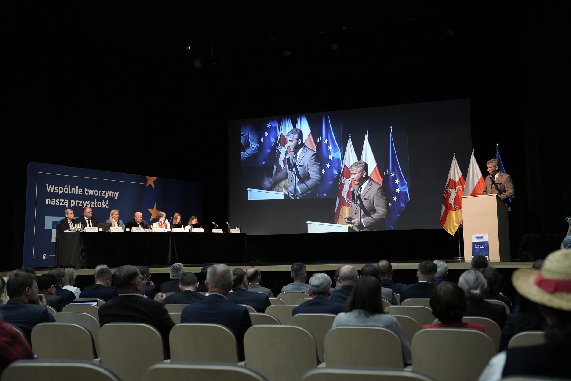 Duża sala konferencyjna z ogromnym ekranem. Na miejscach siedzących znajduje się kilkadziesiąt osób a na scenie obok ekranu wyświetlającego siedzą członkowie prezydium KM i przedstawicielki Komisji Europejskiej