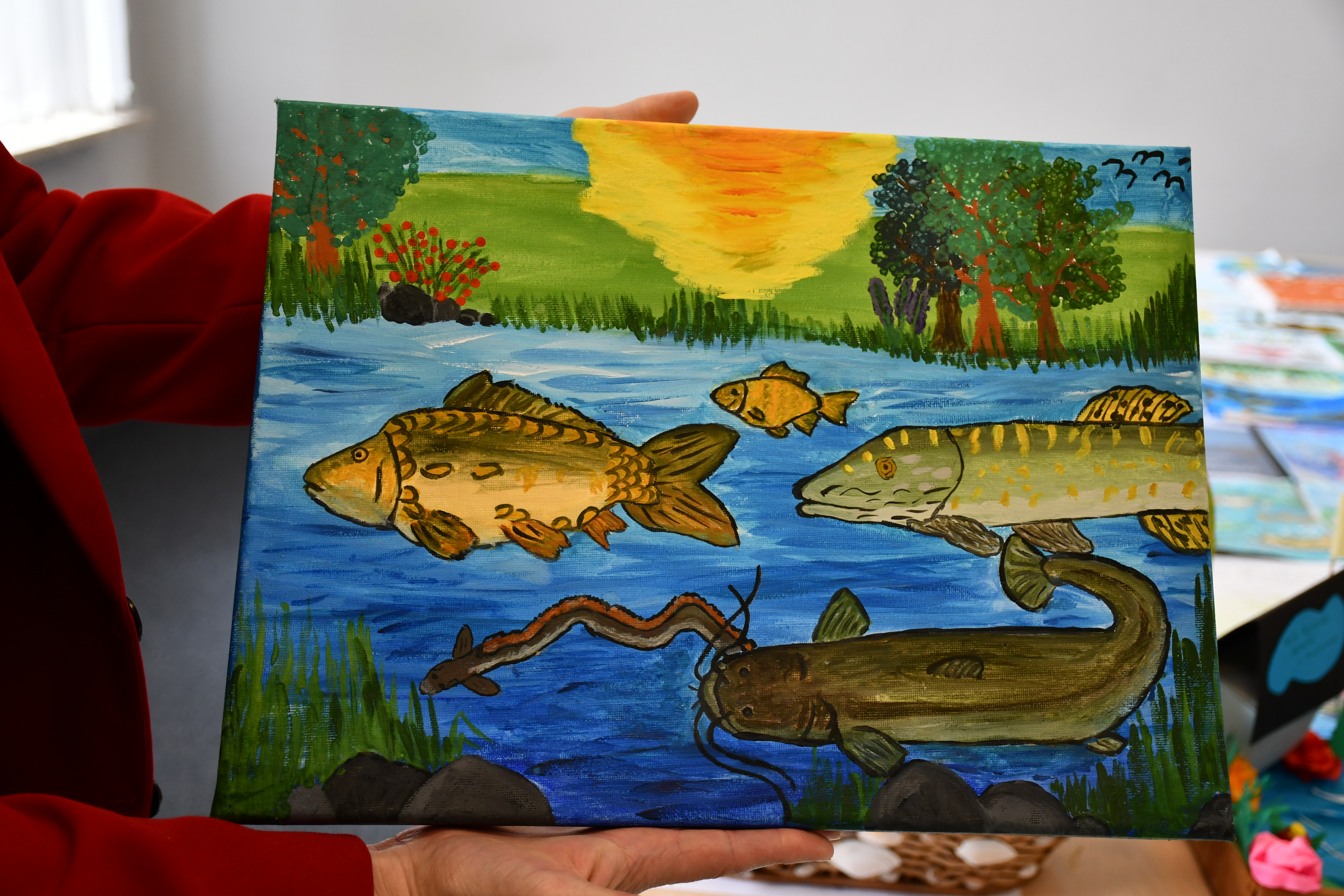 Dłonie trzymające jedną z prac konkursowych, która przedstawia ryby pływające w rzece.