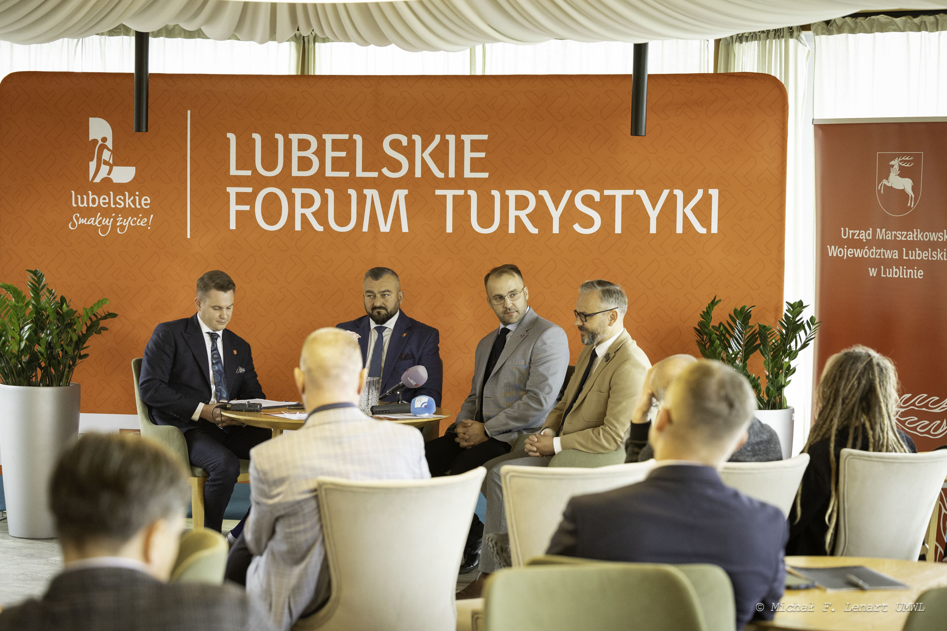 Pod ścianką z napisem Lubelskie forum turystyki ustawiony jest stolik przy którym siedzi czterech mężczyzn. Na pierwszym planie widać pierwsze rzędy uczestników konferencji prasowej