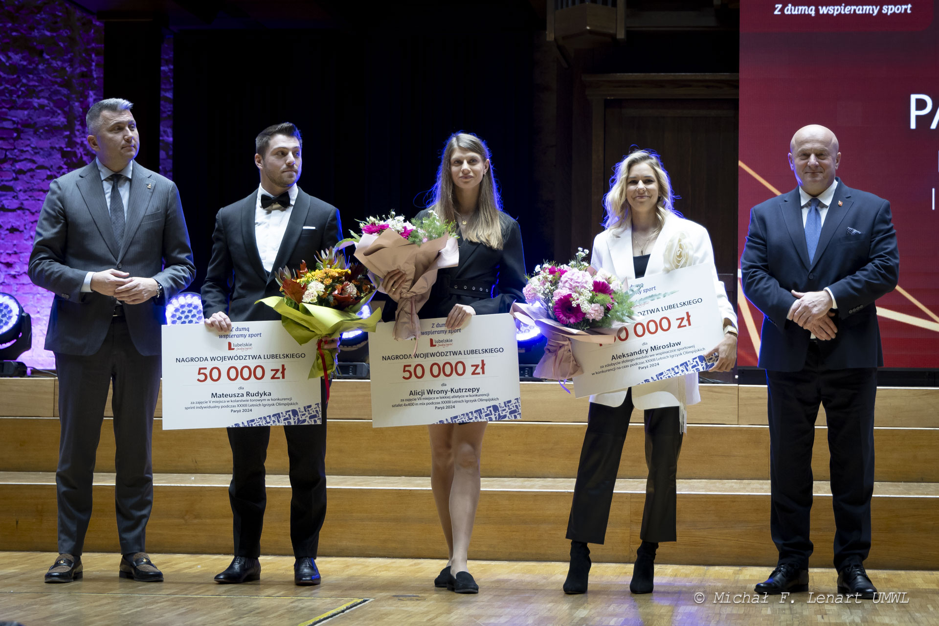 Marszałek Jarosław Stawiarski w towrzystwie tójki olimpijczyków, wszyscy stoją na scenie a sportowcy dodatkowo trzymają w rekach kwiaty i czeki