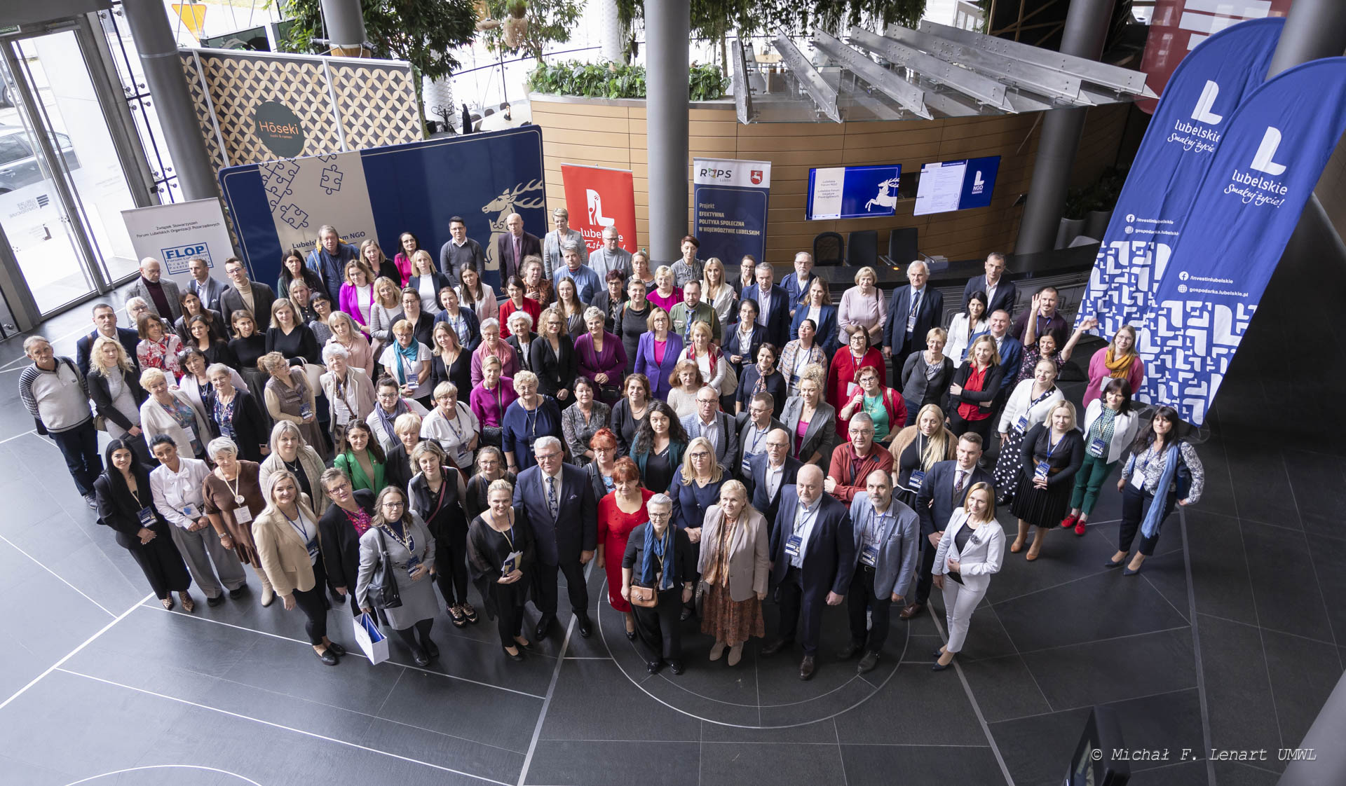 Ujęcie z góry, na sali rejestracyjnej ustawionych jest około pięćdziesięciu osób które wspólnie pozują do zdjęcia