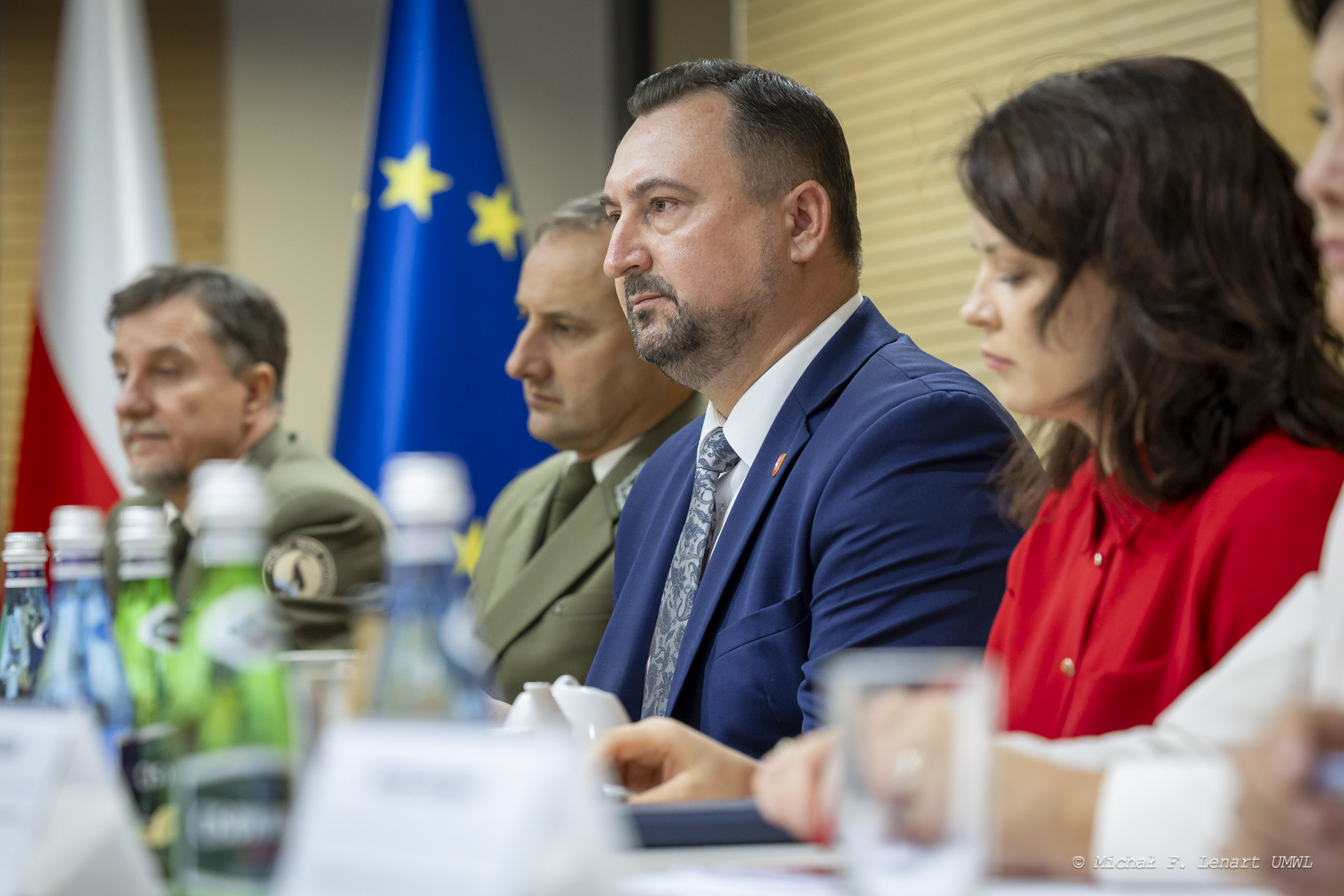 Grupa ludzi siedzących przy stole, z flagami Polski i Unii Europejskiej w tle.