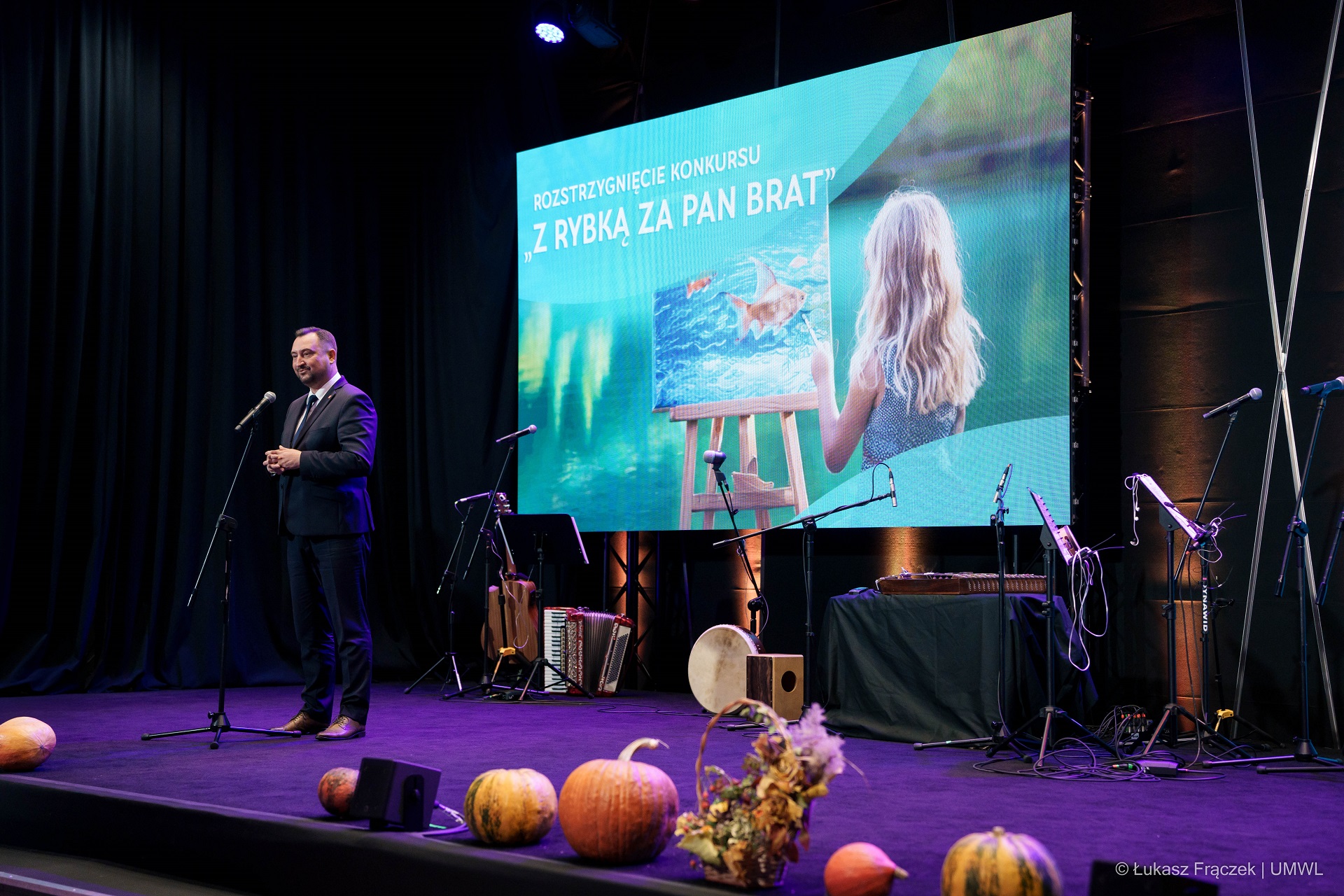 Wicemarszałek Marek Wojciechowski stoi na scenie za nim na duży ekranie wyświetla się grafika dziewczynka która maluje obraz z rybką