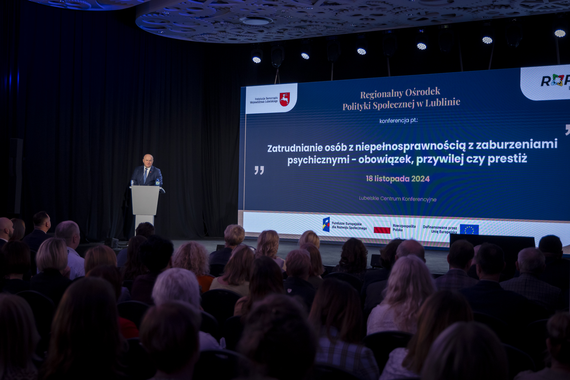 Na pierwszym planie widać pierwsze rzędy publiczności, ktora uczestnicy w konferencji, marszałek Jarosław Stawiarski stoi przy mównicy i zwraca się do zebranych