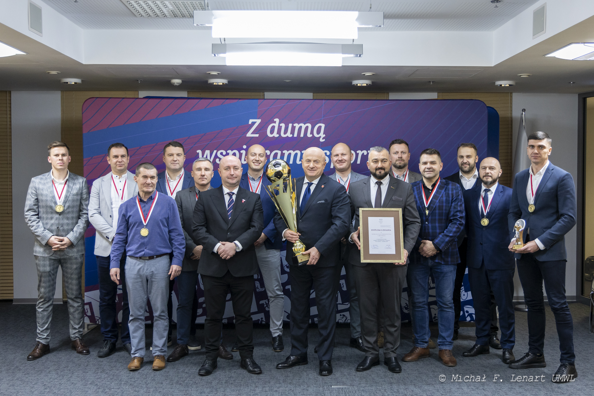 Drużyna urzędu marszałkowskiego wspólnie pozuje do zdjęcia z członkami zarządu w sali konferencyjnej. Członkowie zarządu prezentują zdobyty przez drużynę puchar i dyplom