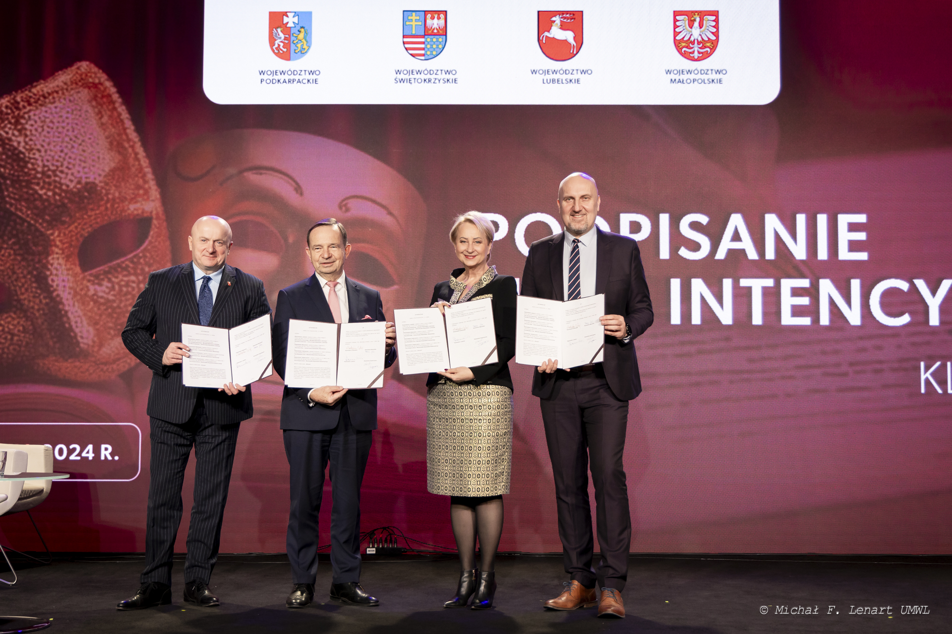 Trzech mężczyzn i jedna kobieta, wszyscy stoją na scenie i prezentują w otwartych teczkach podpisany list intencyjny