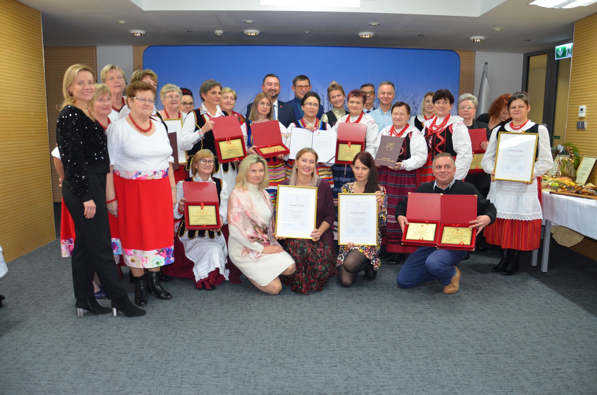 Kilkanaście kobiet w strojach ludowych stoi w sali konferencyjnej i pozuje do zdjęcia wspólnie z wicemarszałkiem Markiem Wojciechowskim
