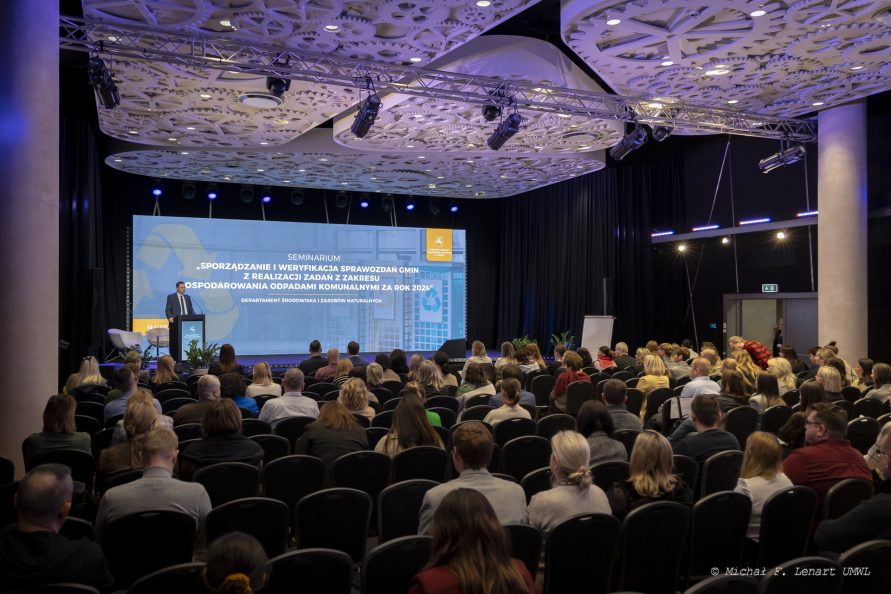Sala konferencyjna widziana z tyłu. Kilkadziesiąt osób siedzących. W tle widok slajdu z tytułem seminarium rozciągającego się na całą ścianę. W głębi po lewej stoi jeden z prelegentów.