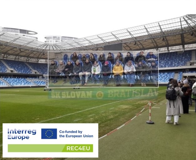 Stadion ŠK Slovan Bratysława, czyli wizyta studyjna w drugim dniu spotkania partnerów w Bratysławie. Partnerzy stoją na stadionie omijając murawę.