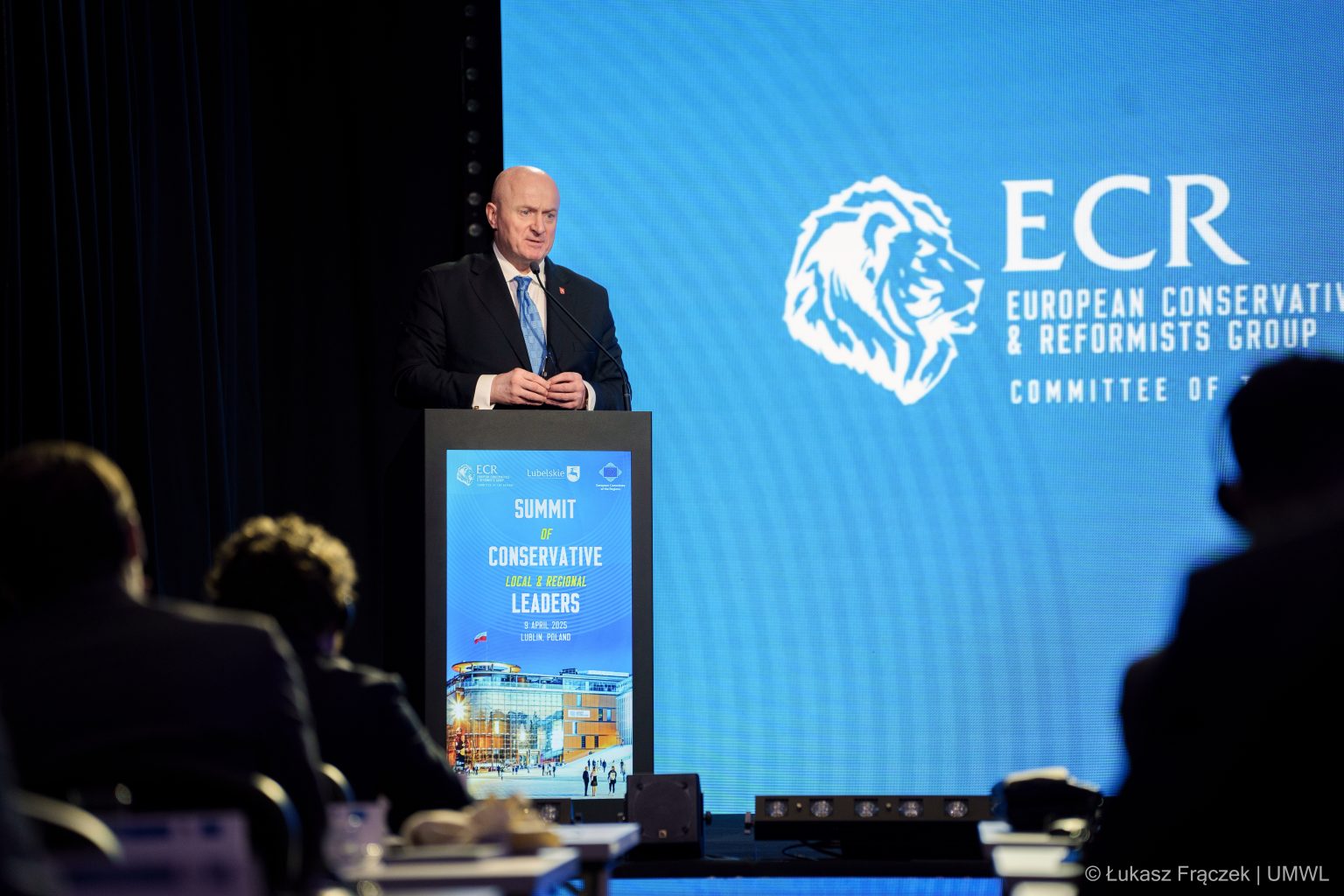 Marszałek Jarosław Stawiarski przemawia ze sceny. W tle logo przedstawiające głowę lwa i napis ECR - European Conservatives and Reformist Group, Committee of Regions (Europejskie Ugrupowanie Konserwatystów i Reformatorów, Komitet Regionów)