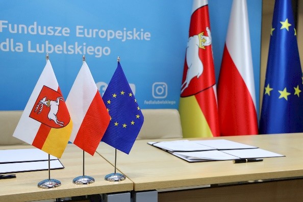 Obraz przedstawia grafikę na której widnieją 3 flagi: Województwa Lubelskiego, Polski oraz Unii Europejskiej, w tle na niebieskim tle napis" Fundusze Europejskie dla Lubelskiego.