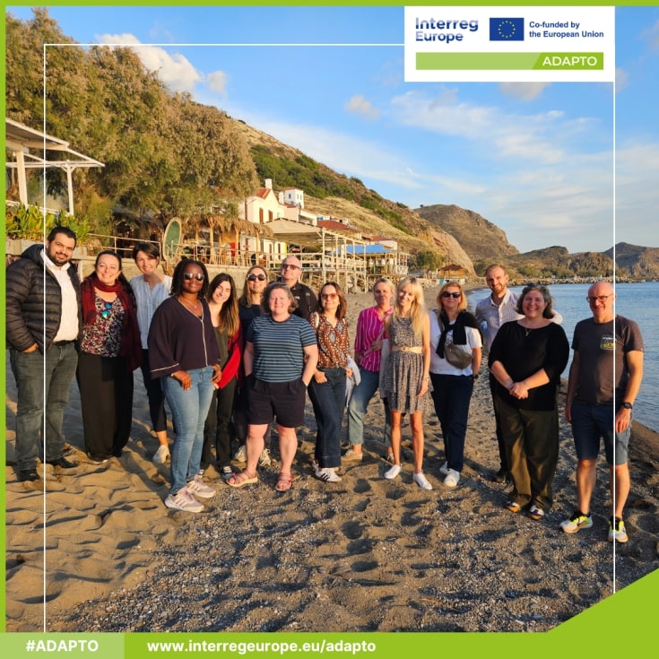 Partnerzy ADAPTO nad Morzem Egejskim. widać kawałek plaży i promienie Słońca. Wszyscy stoją przodem do obiektywu.