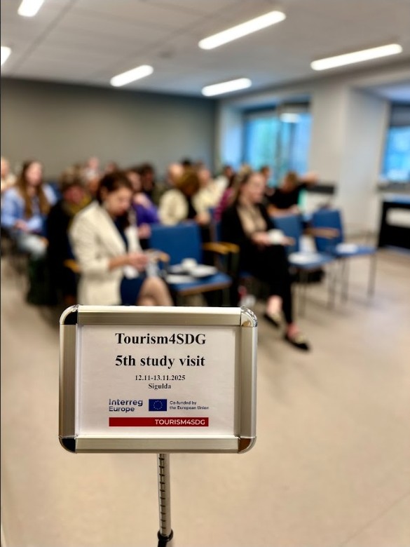 Tablica informacyjna z logiem projektu, w tle uczestnicy słuchają prelekcji.